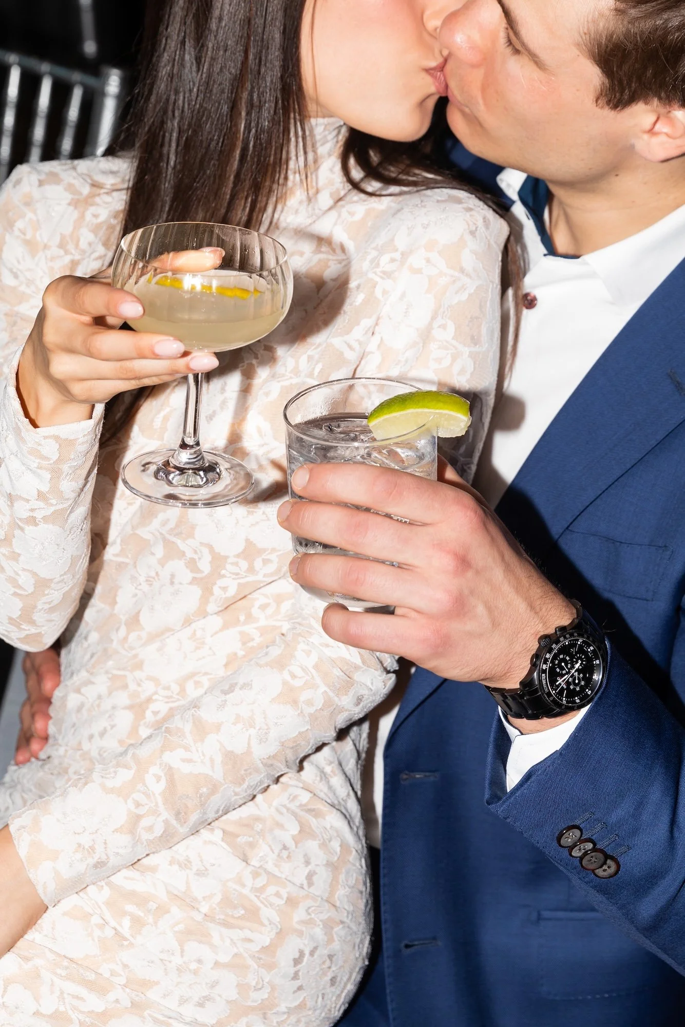 A couple at a social gathering sharing a kiss, with the woman holding a cocktail glass and the man holding a glass with lime, both dressed in formal attire.