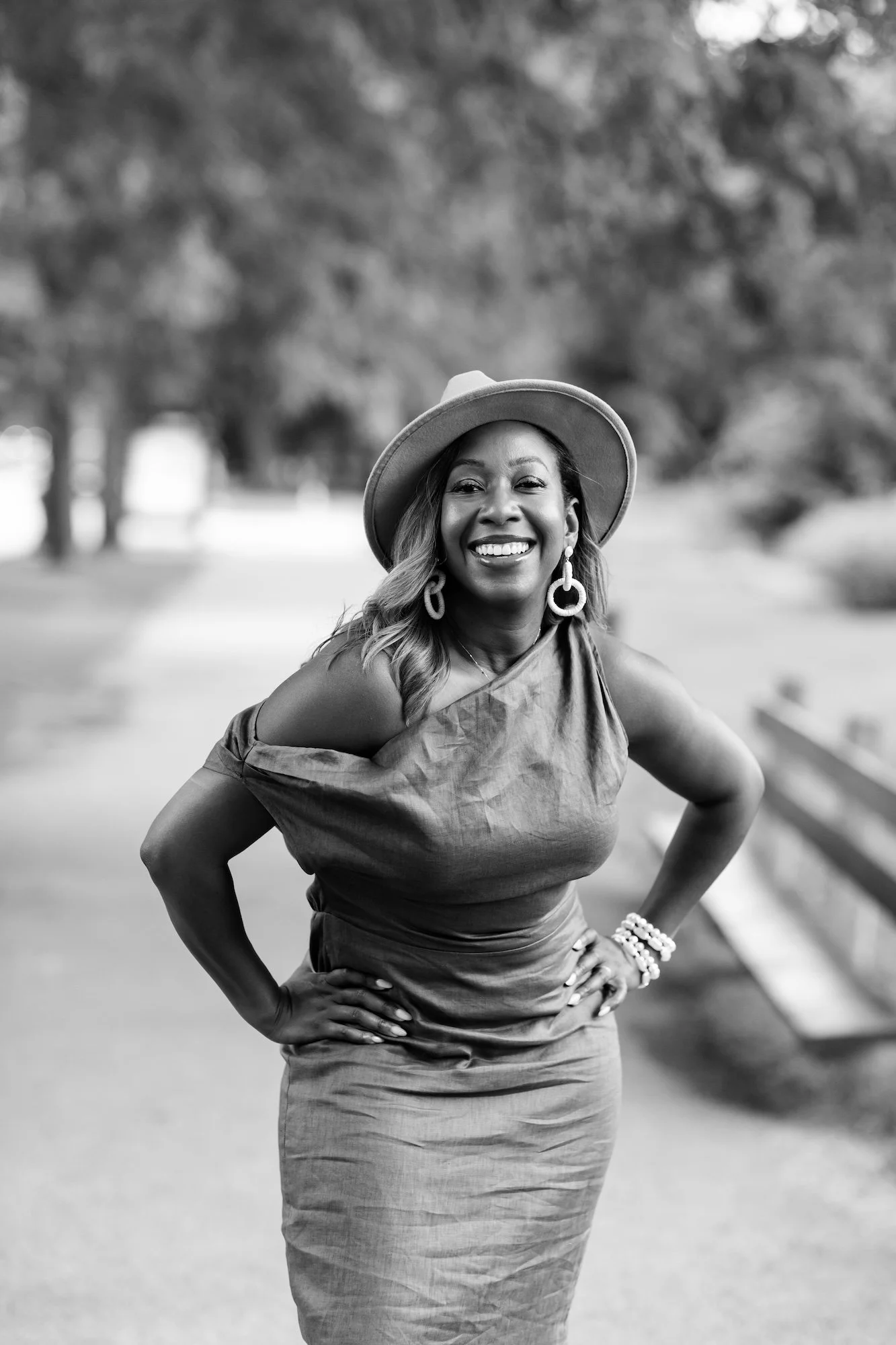 A smiling woman in a dress and wide-brimmed hat standing outdoors on a pathway with trees in the background.
