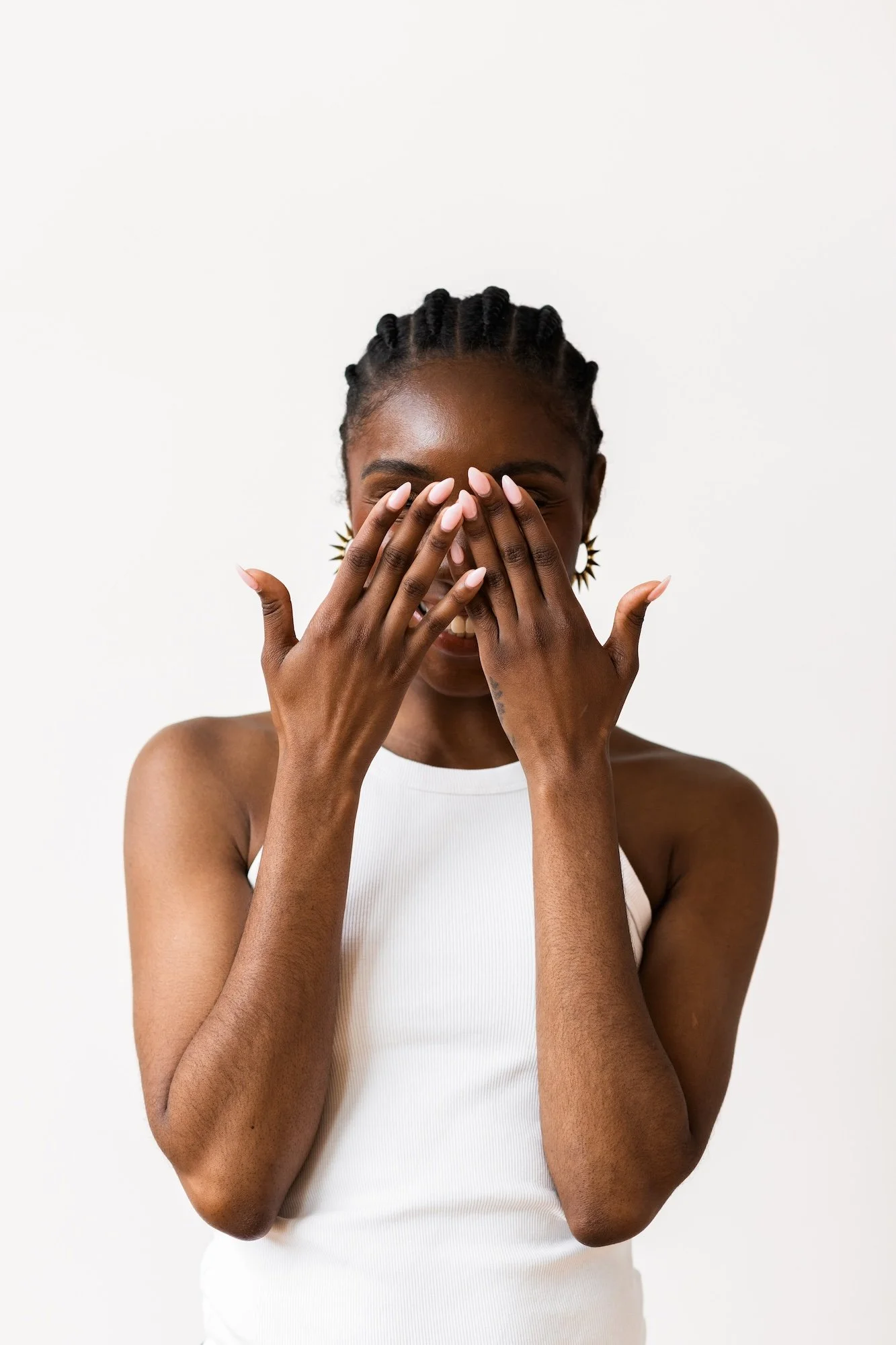 A woman with braided hair, wearing gold earrings and a white sleeveless top, is covering her face with her hands and smiling, standing against a plain white background.