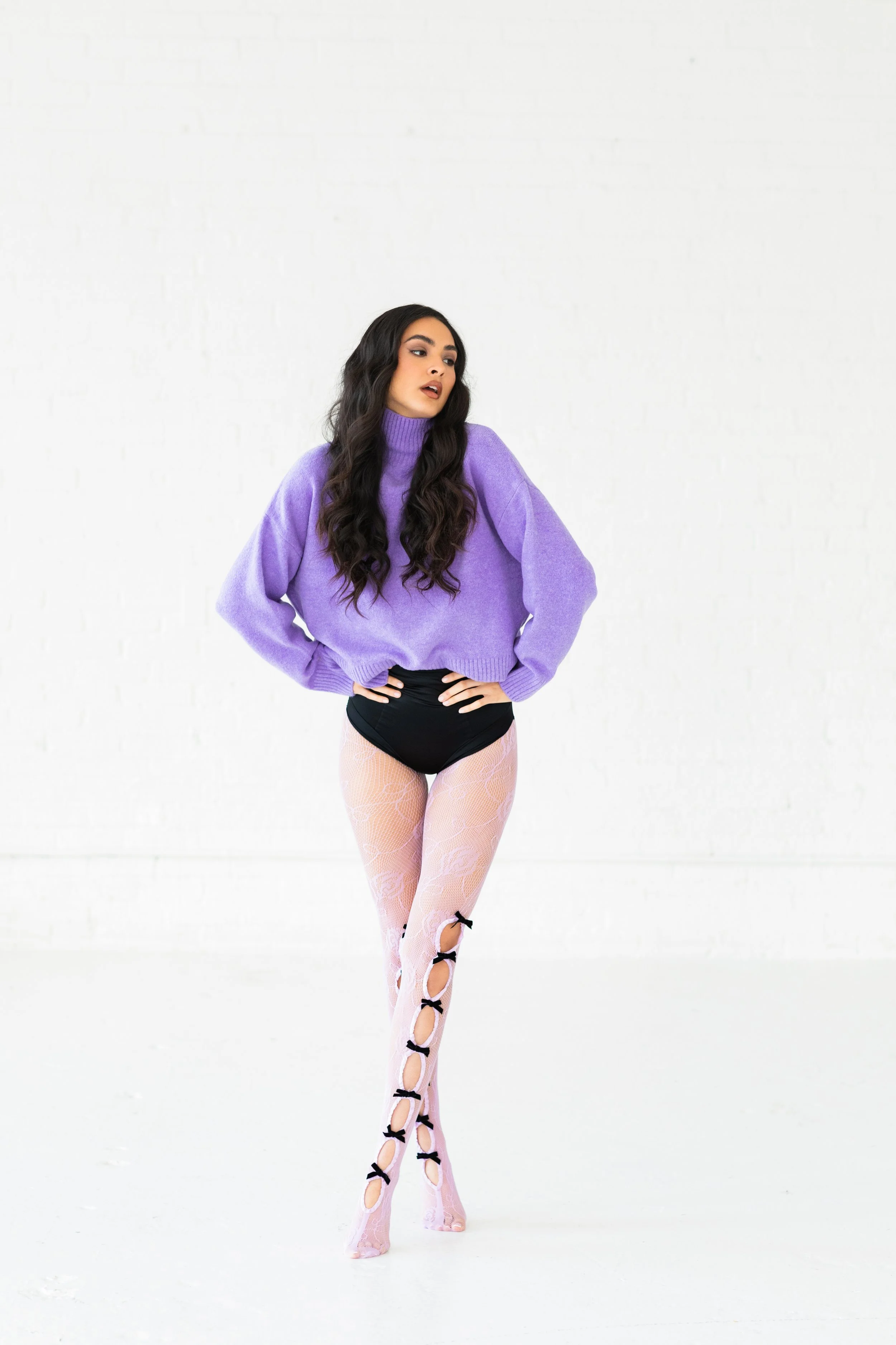 Woman with long dark hair wearing a purple sweater, black underwear, and pink lace thigh-high stockings with black bows, standing in a white space.