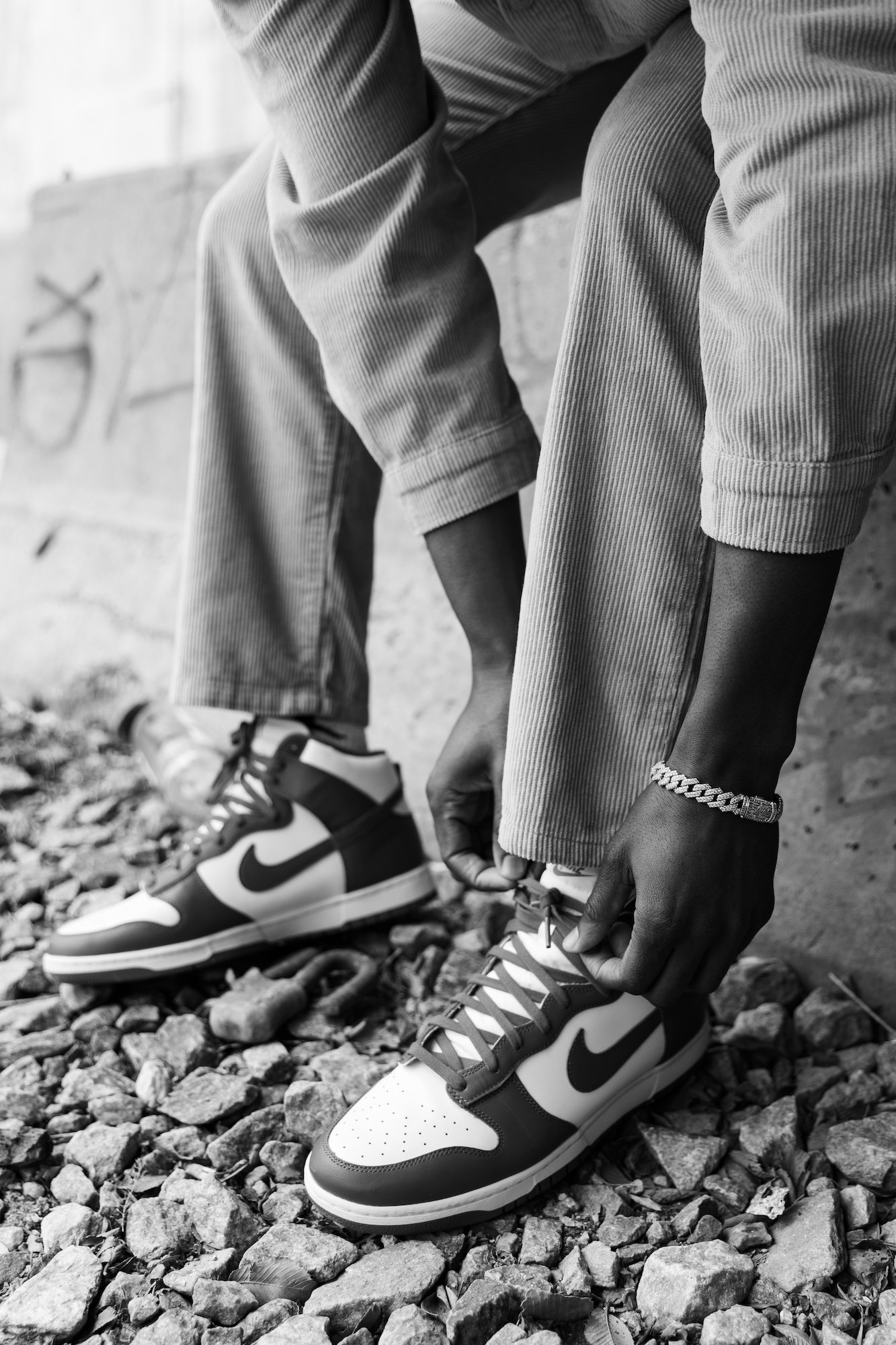 A person wearing striped pants is tying the laces of sneakers with Nike swooshes on a gravel surface, with graffiti in the background.