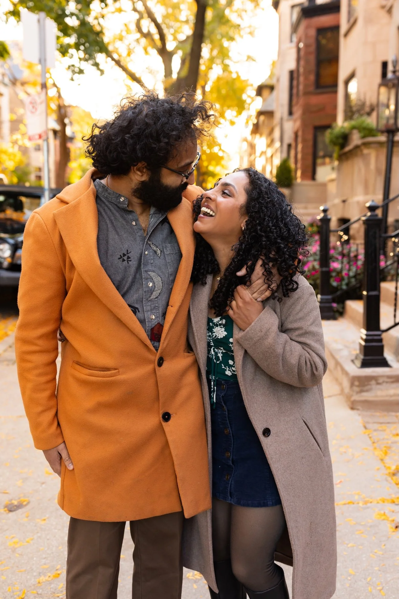 A couple standing close together on a city sidewalk, smiling and looking into each other's eyes on a fall day.