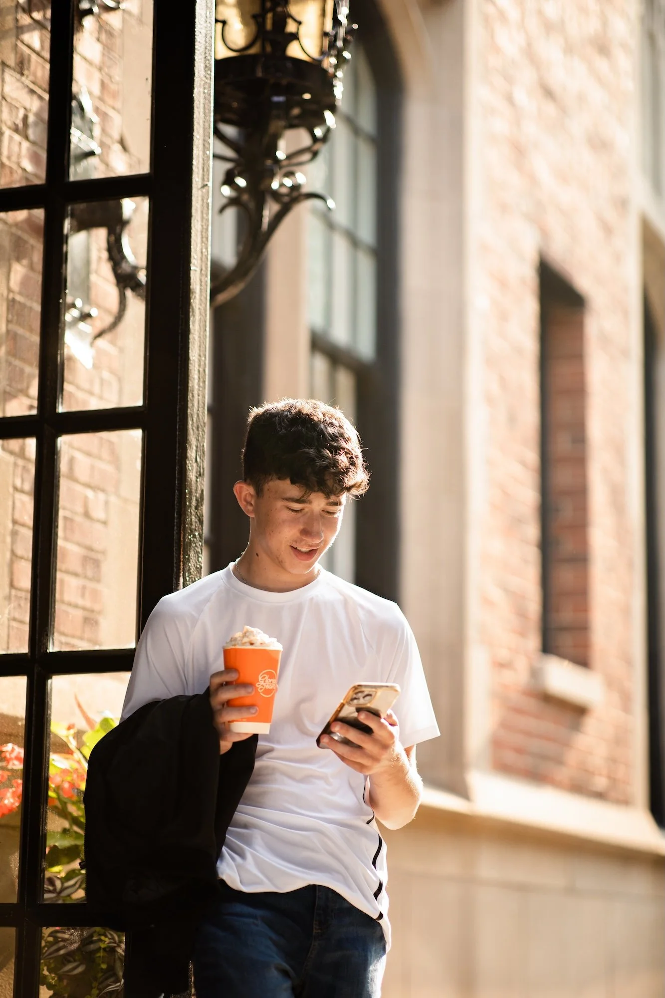 A teenage boy stands outside, looking at his smartphone while holding a cup of ice cream in his other hand. He is wearing a white t-shirt and has a black jacket draped over his shoulder. The background features brick walls, large windows, and outdoor lighting fixtures.