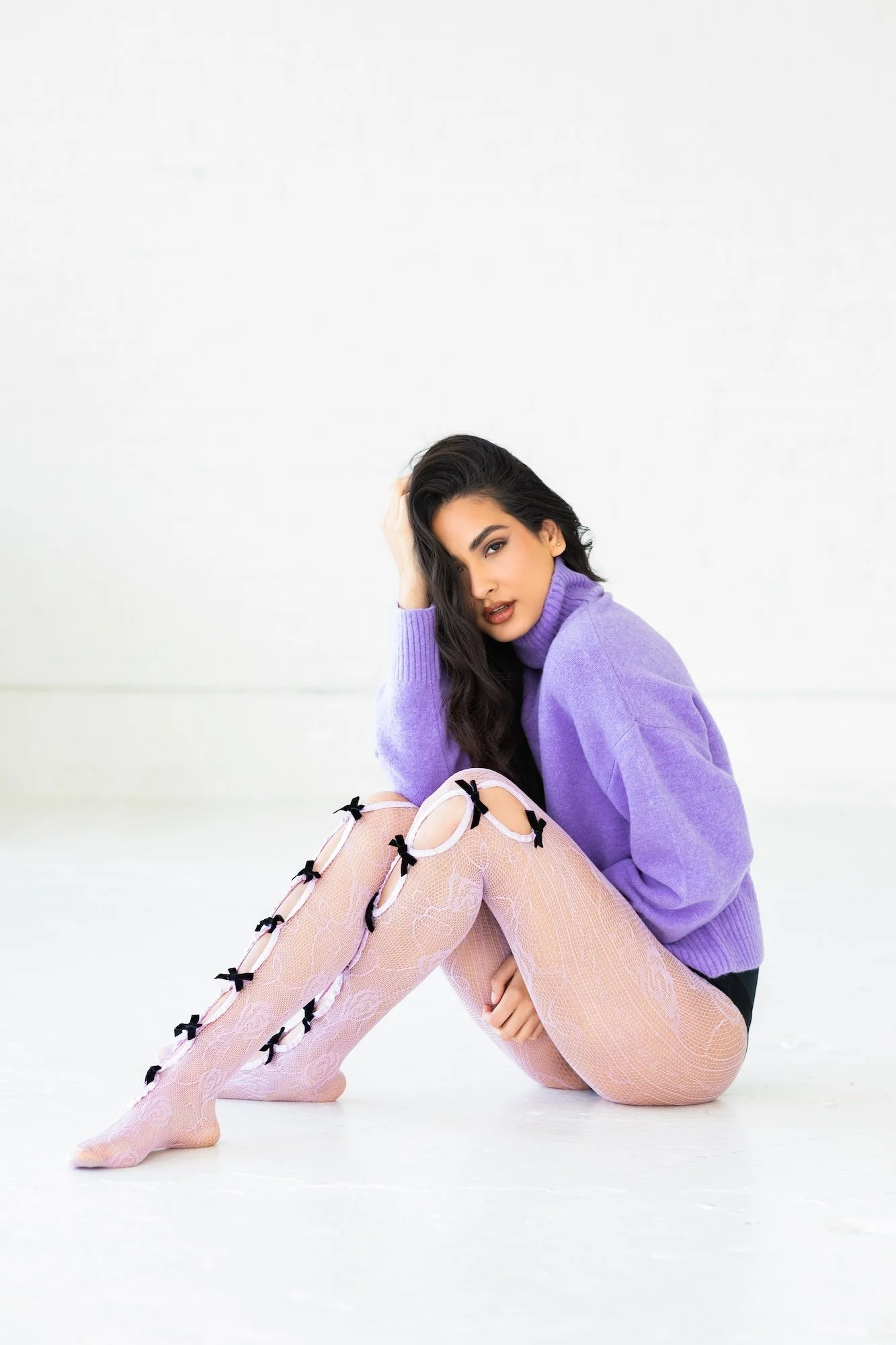 A woman with long black hair, wearing a purple sweater and pink lace thigh-high stockings with black bows, sitting on the floor with white background, in a relaxed pose.