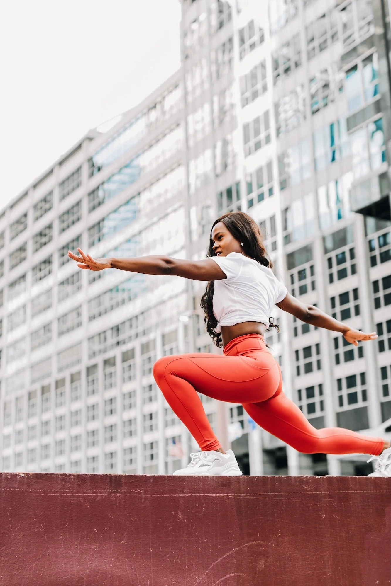 A woman in a white crop top and coral leggings performing a yoga pose outdoors on a red platform, with tall modern buildings in the background.