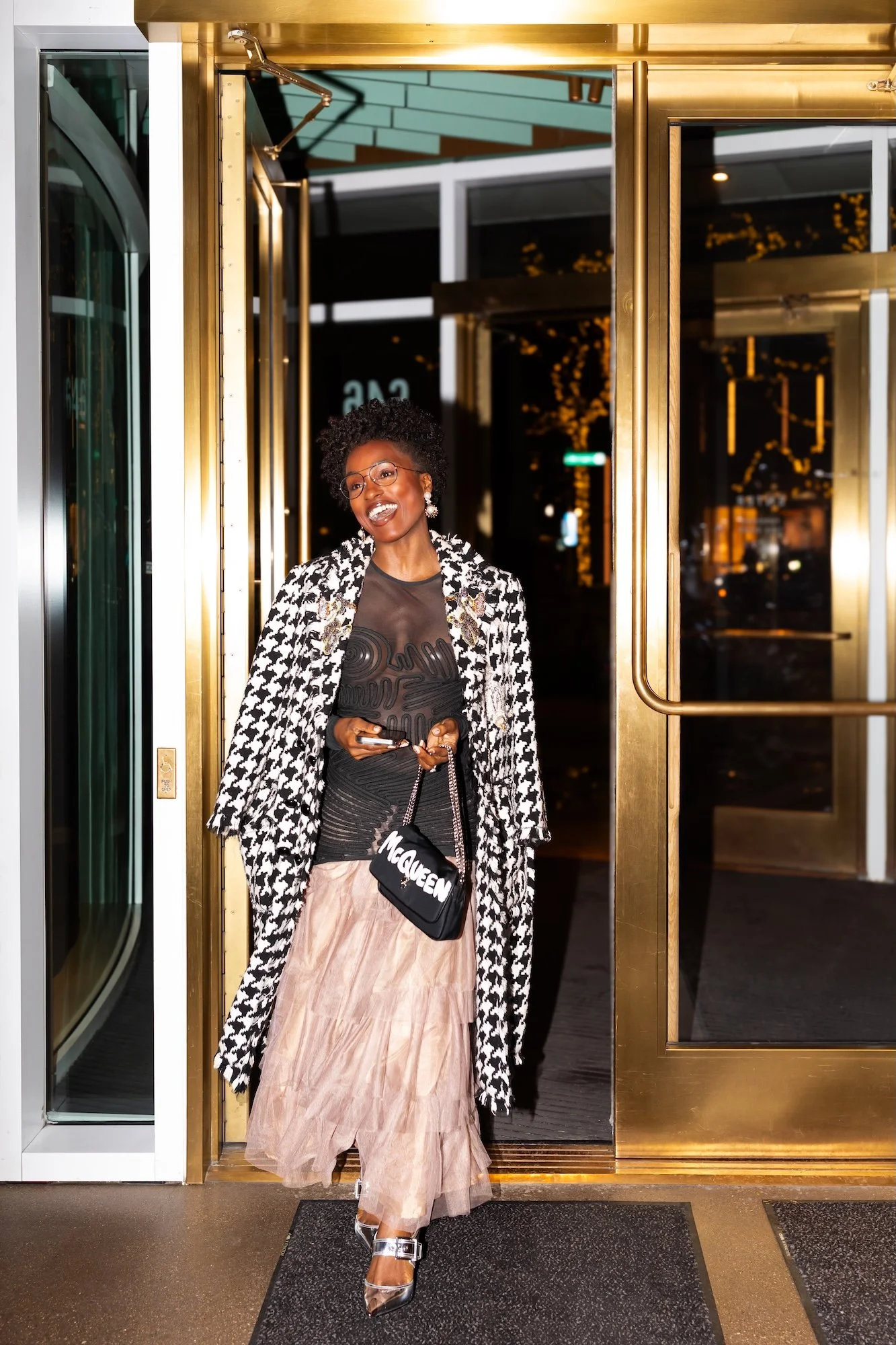 A woman dressed in black and beige, wearing glasses and jewelry, exiting a building with large glass doors and golden frames, smiling and holding a phone and small black McQueen purse.