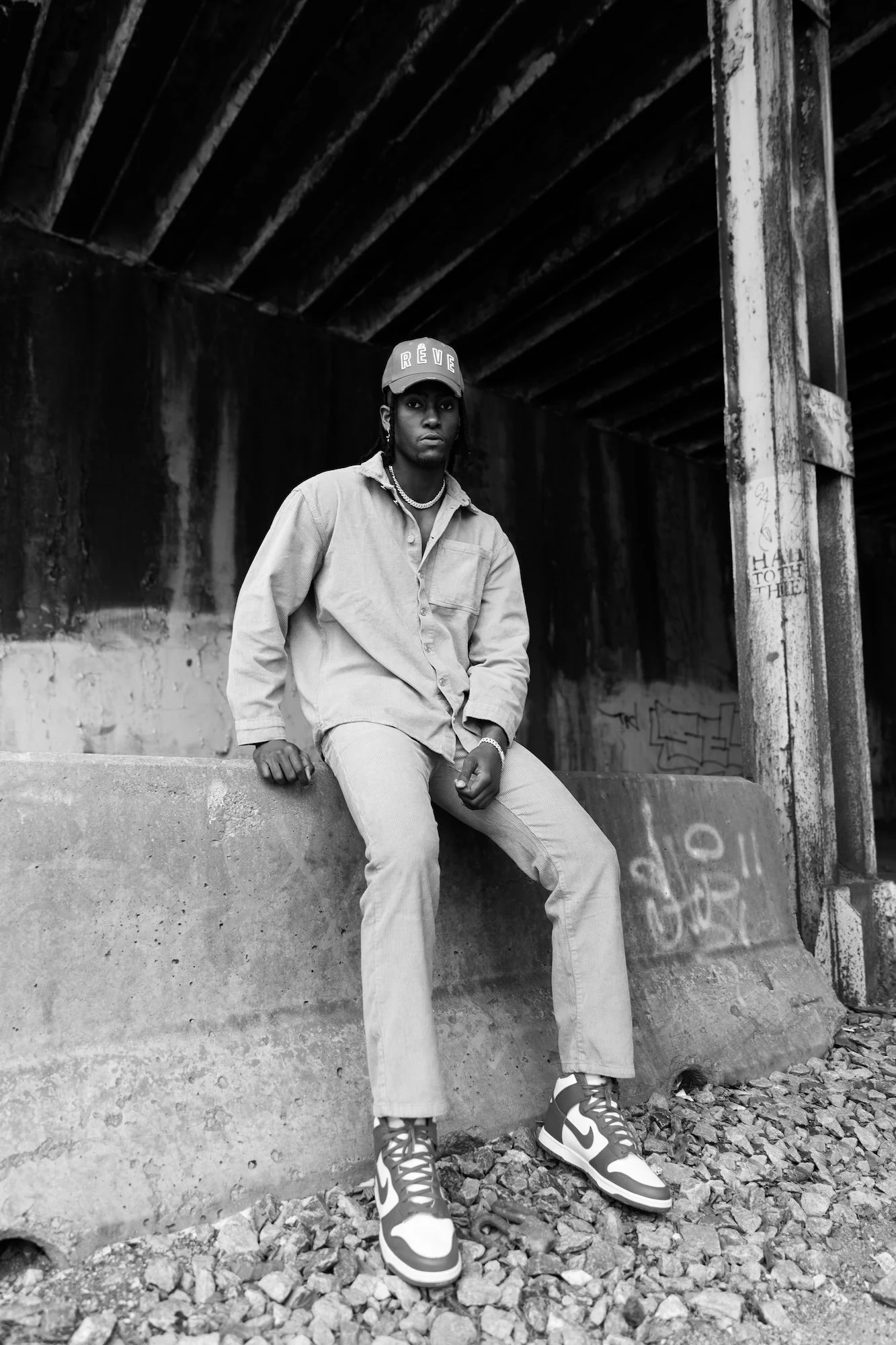 A young man sitting on a concrete ledge under a bridge, dressed in casual streetwear, wearing a cap, jewelry, and sneakers, in black and white photography.