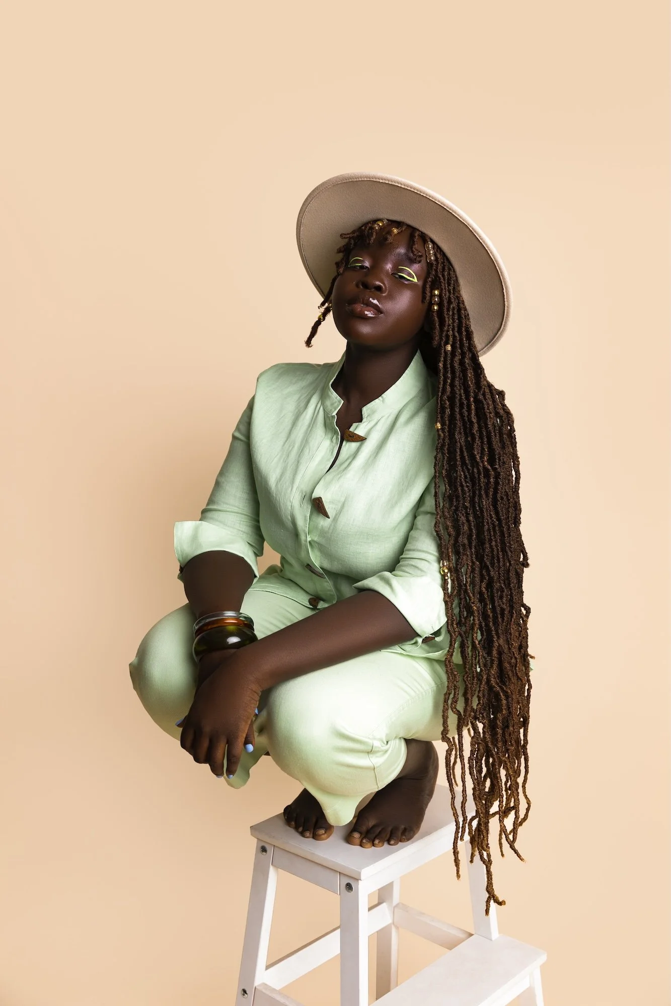 A woman with long dreadlocks, wearing a beige hat and light green outfit with rolled-up sleeves, sitting on a white stool against a beige background.
