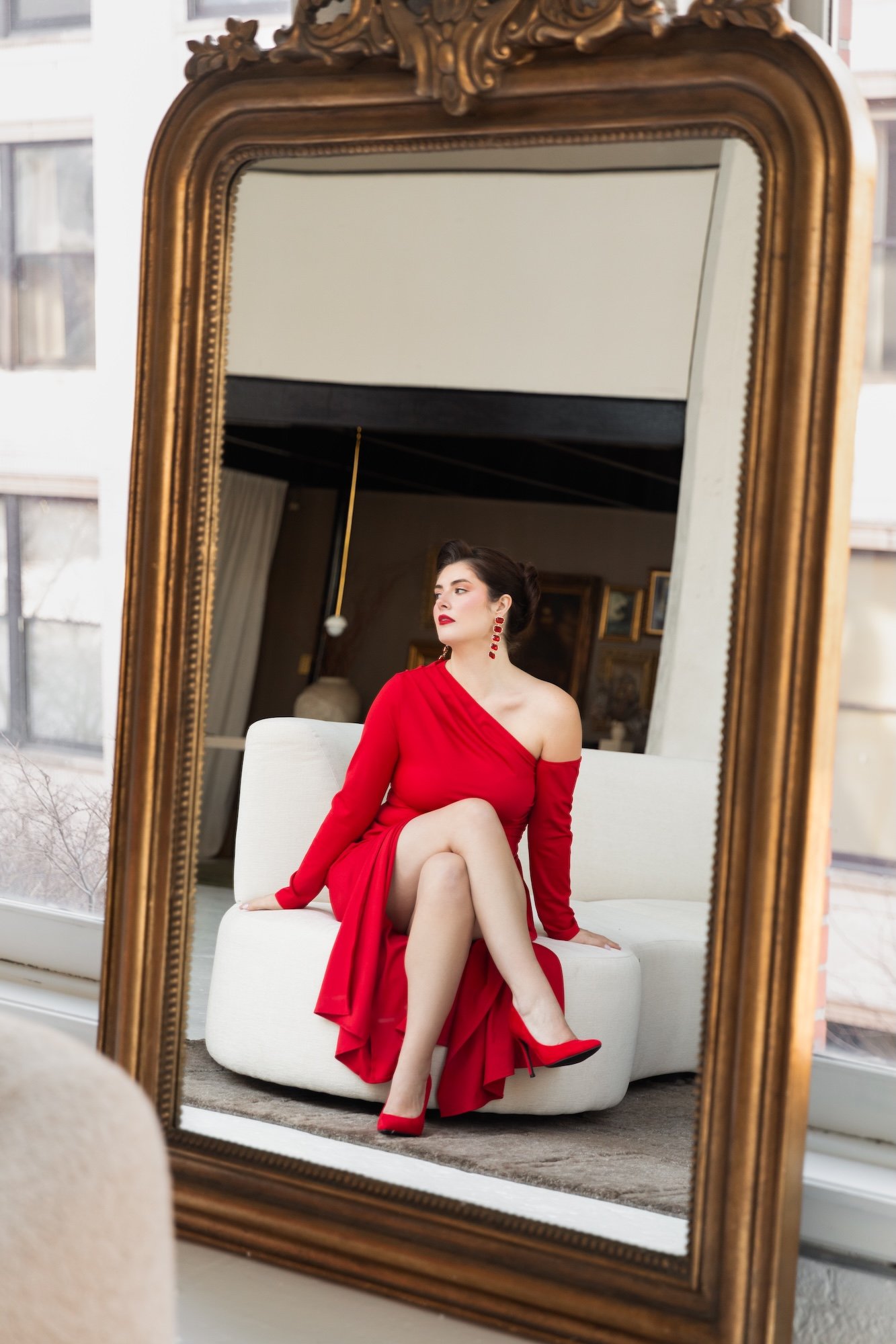 A woman in a red dress and matching red high heels sitting on a white sofa, looking to the side, seen through a large ornate mirror with a gold frame.