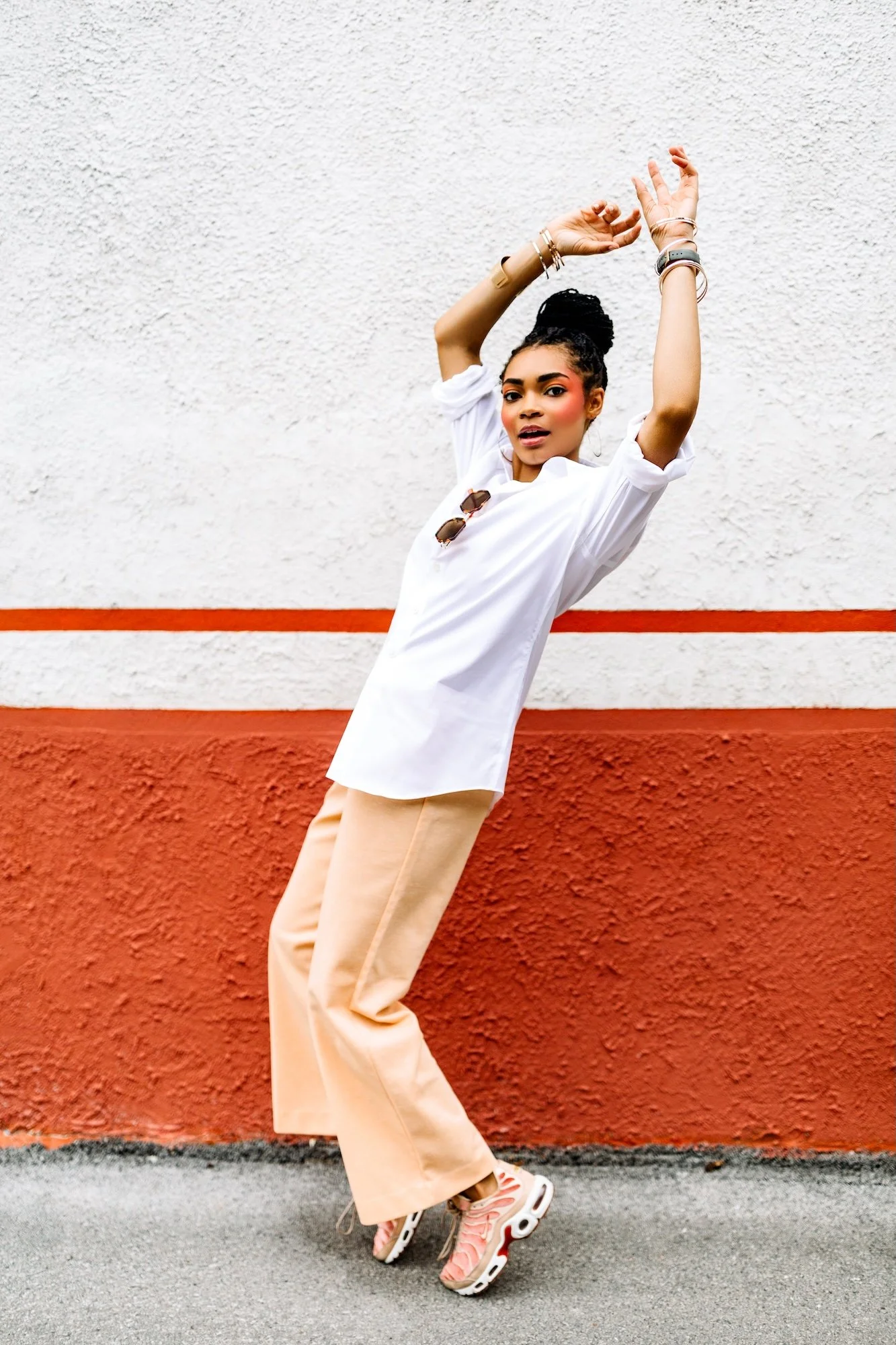 A young woman with dark hair in a bun, wearing a white button-down shirt, beige pants, and trendy sneakers, standing against a textured wall with a white upper half and red lower half, striking a pose with her arms raised and one leg bent.