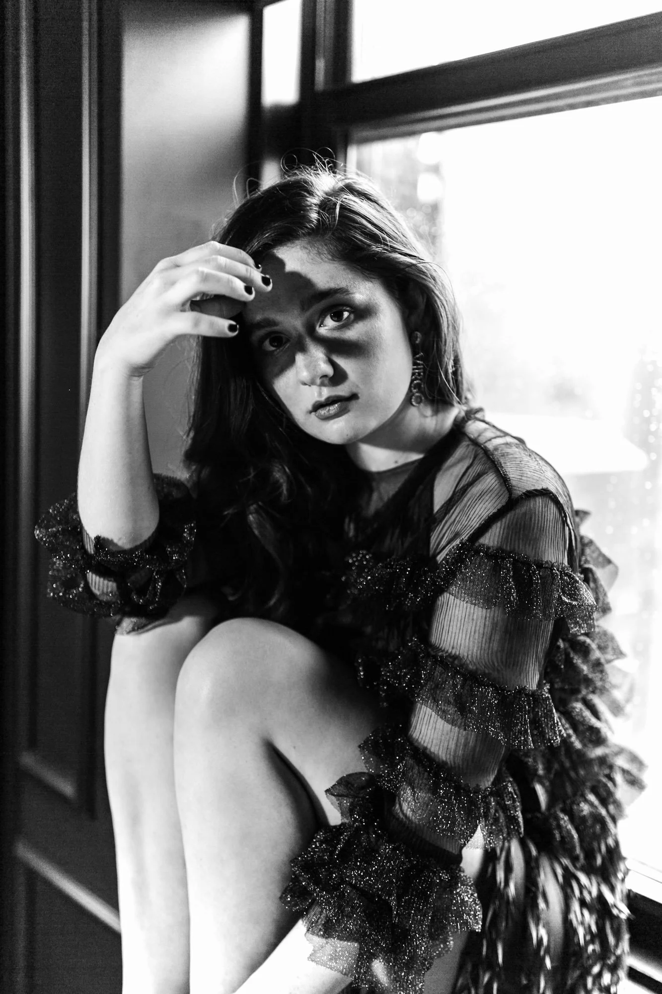 A black-and-white photo of a young woman sitting by a window, with her hand partly covering her face. She wears a textured, ruffled, semi-transparent blouse with sparkling details and has long, wavy hair.