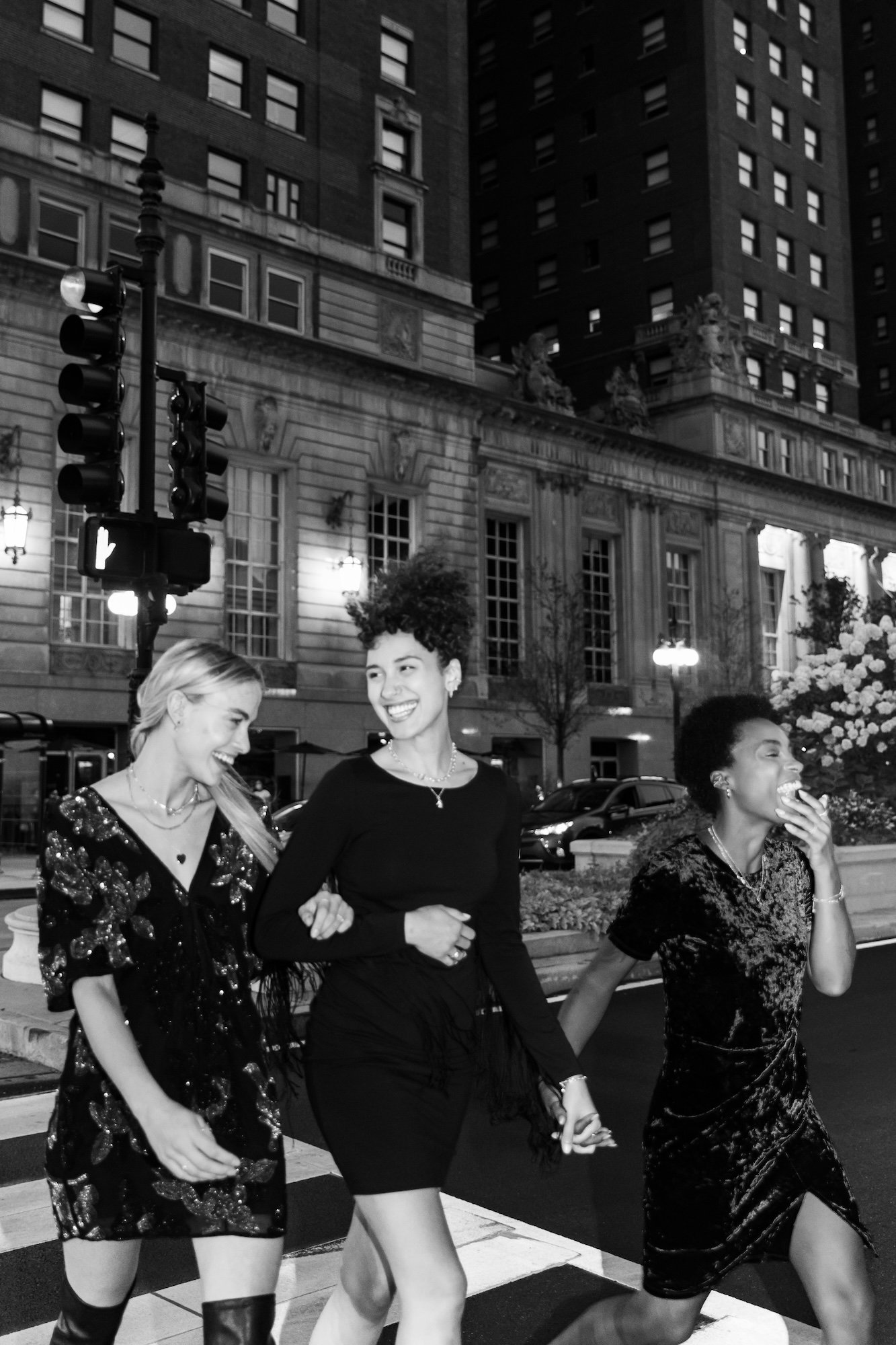Three women are crossing a city street at night, laughing and holding hands. The background features a historic building, traffic lights, and parked cars.
