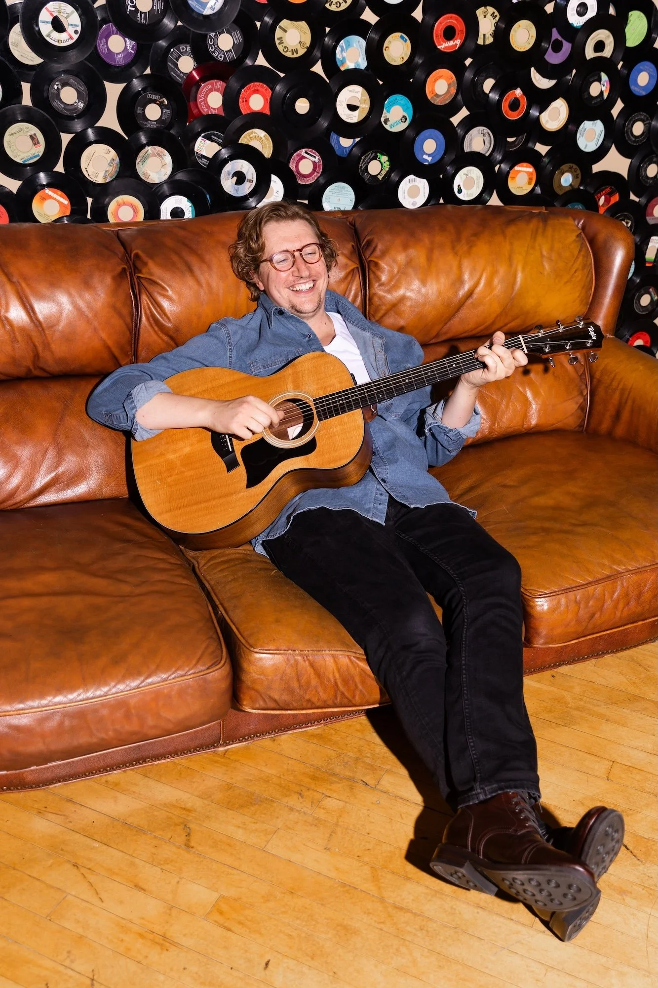 A young man with glasses and curly hair, smiling and playing an acoustic guitar while sitting on a brown leather couch. The background features a wall decorated with vinyl records.