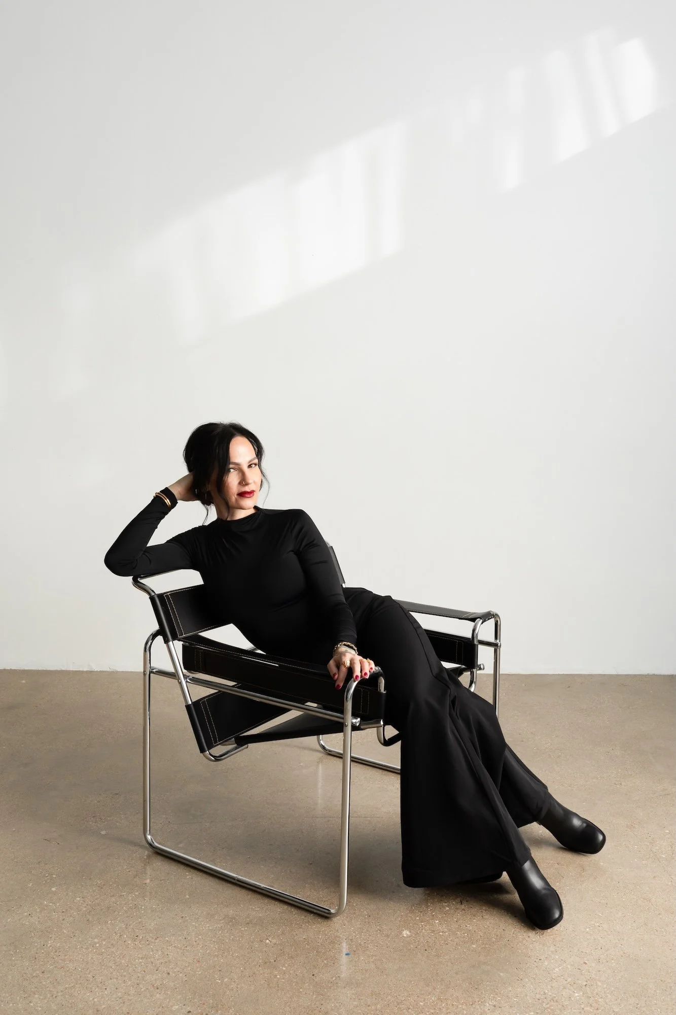 A woman with black hair and red lipstick, wearing a black long-sleeve top and black pants, sitting on a modern black and chrome chair in a minimalist space with a white wall and concrete floor.