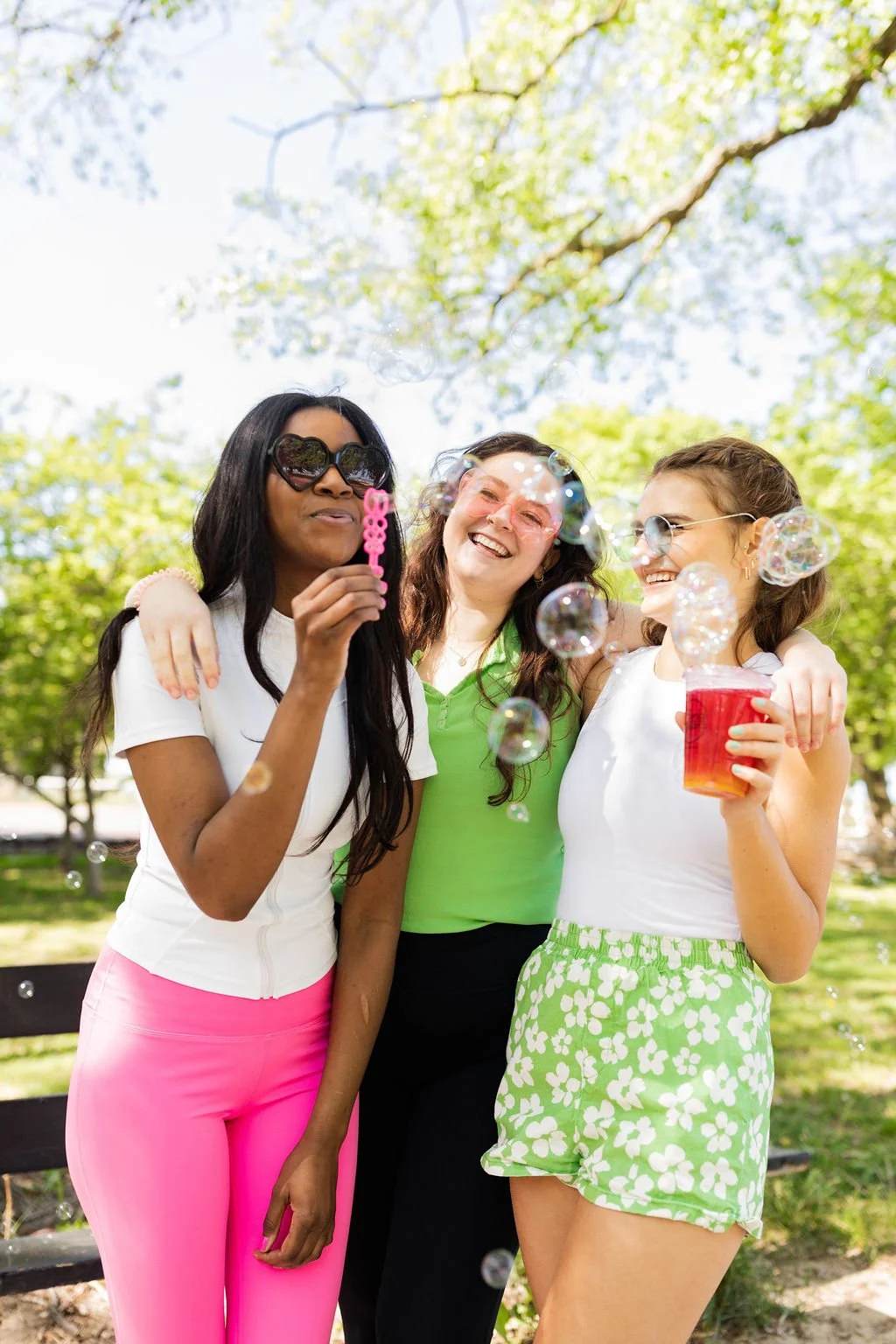 Three women outdoors in a park, smiling and enjoying bubbles, with one blowing bubbles and wearing colorful sunglasses and bright clothing.