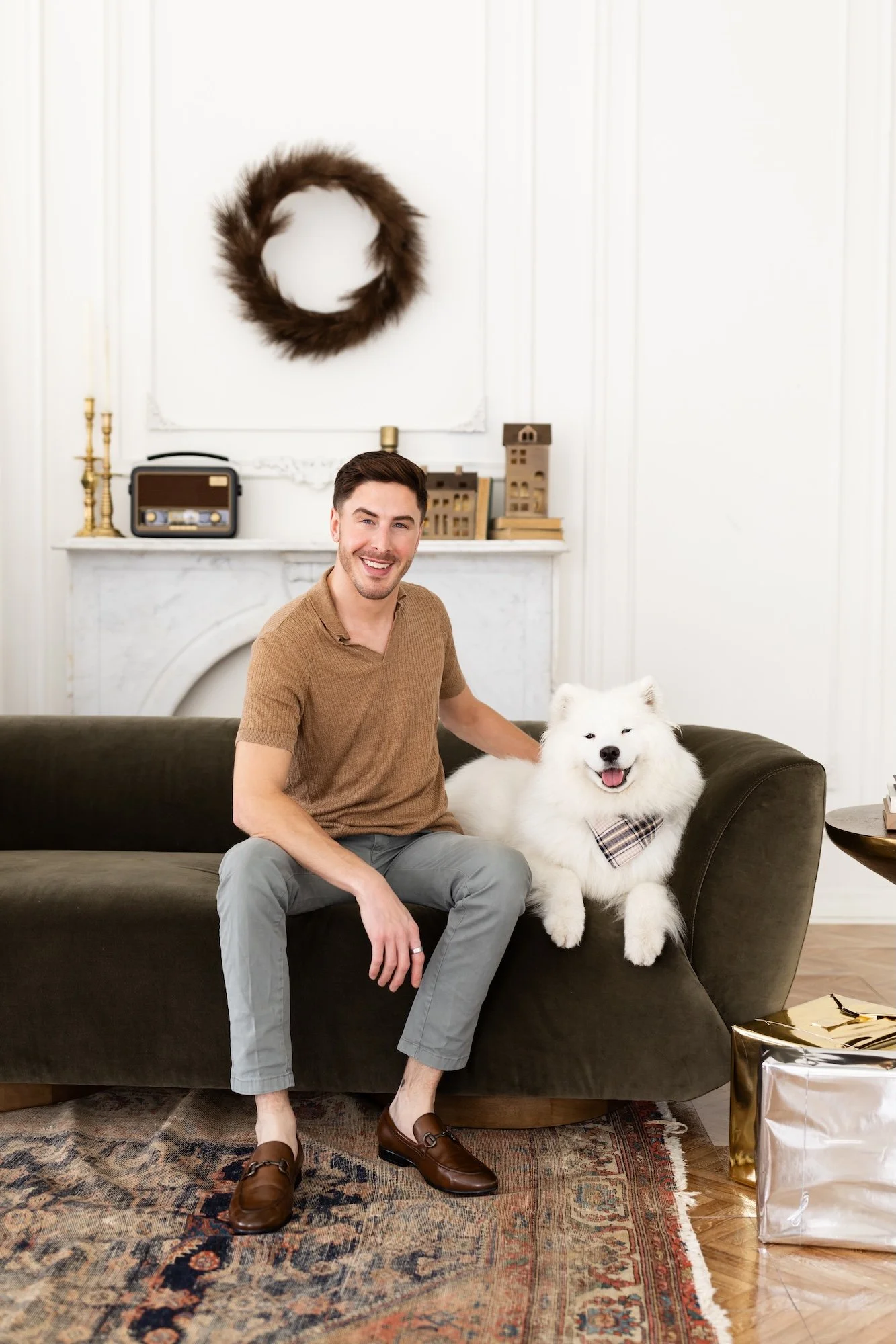 A smiling man sitting on a dark green sofa next to a fluffy white dog with a plaid bandana. The room has white walls, a marble fireplace, and a gold gift box on the floor.