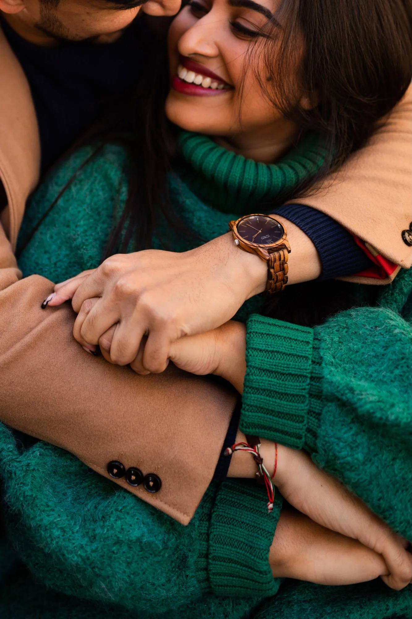 Two people hugging: one with a wooden watch, wearing a green sweater and a beige coat, smiling, and the other with a black shirt and a red bracelet, also hugging.