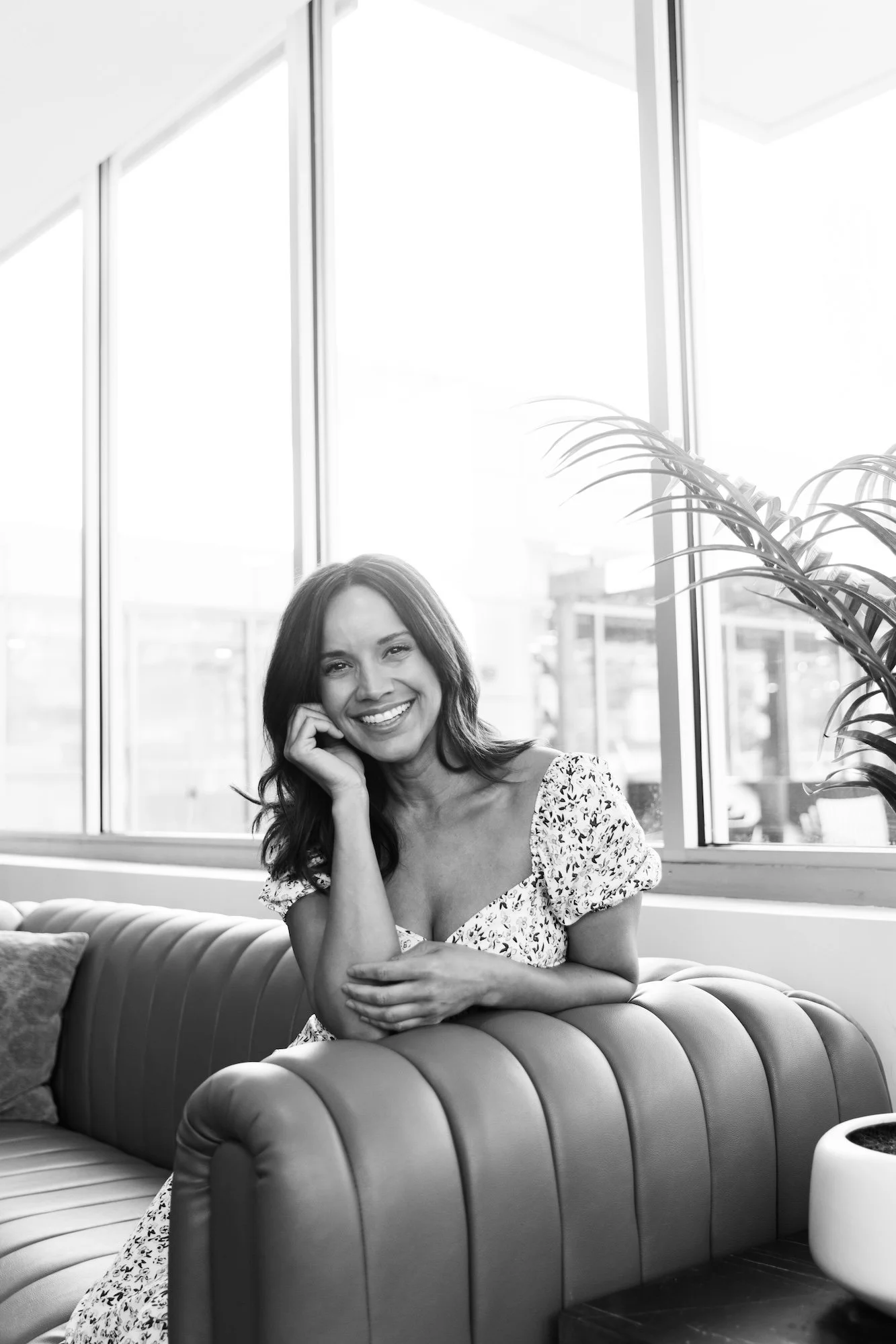 Black and white photo of a smiling woman sitting on a leather sofa, resting her head on her hand, with large windows and a plant in the background.