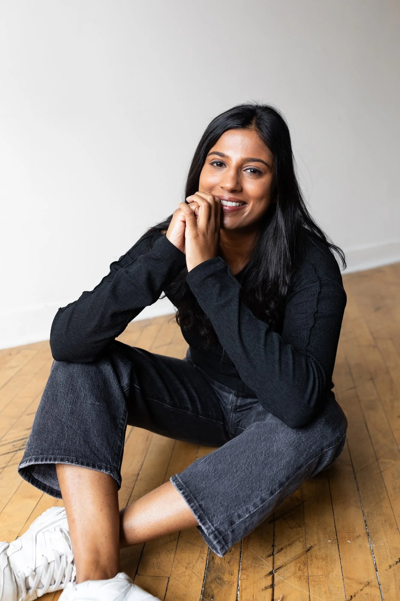 A woman with long black hair sitting on a wooden floor, smiling at the camera, wearing a black long-sleeve shirt, black jeans, and white sneakers, with a plain white wall in the background.