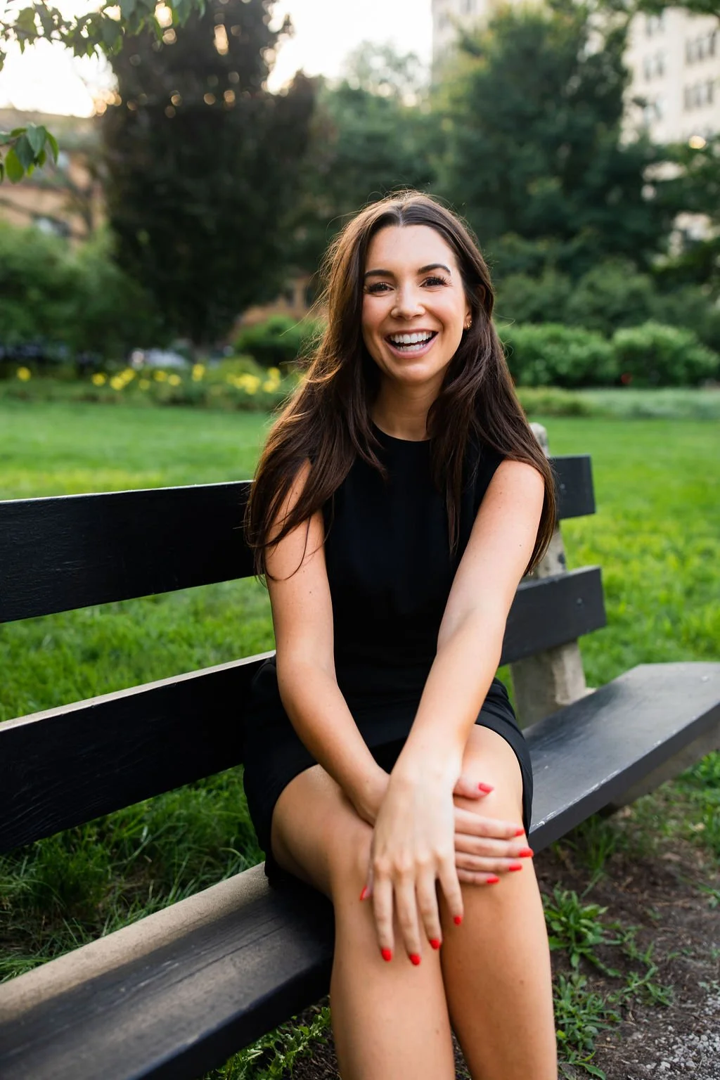 A young woman with long dark hair, wearing a black dress, sitting on a park bench and smiling broadly at the camera.