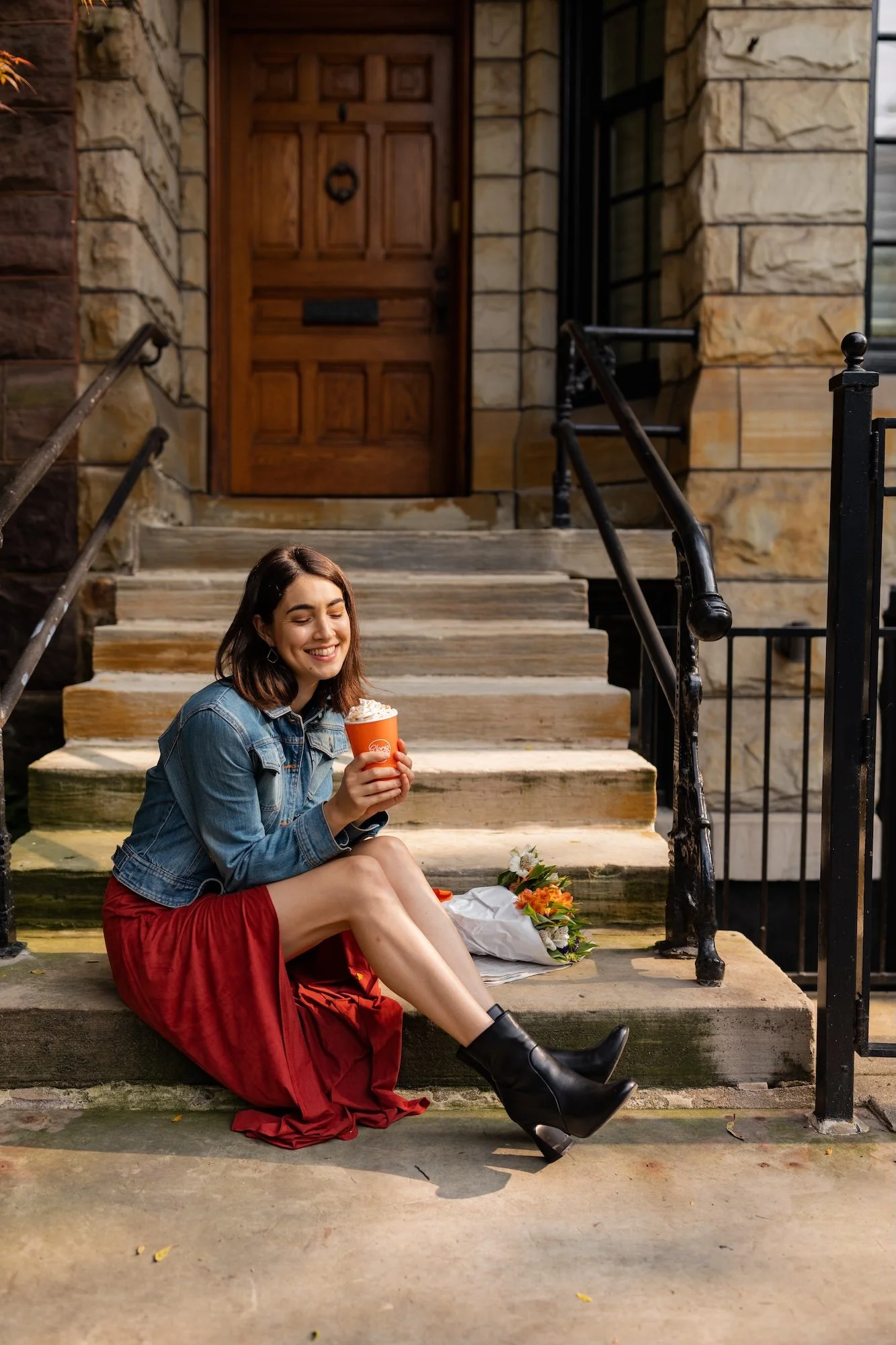 A young woman sitting on the steps of a house, holding a cup of whipped cream, smiling with her eyes closed. She is wearing a denim jacket, red skirt, and black ankle boots. There is a bouquet of flowers on the ground beside her.