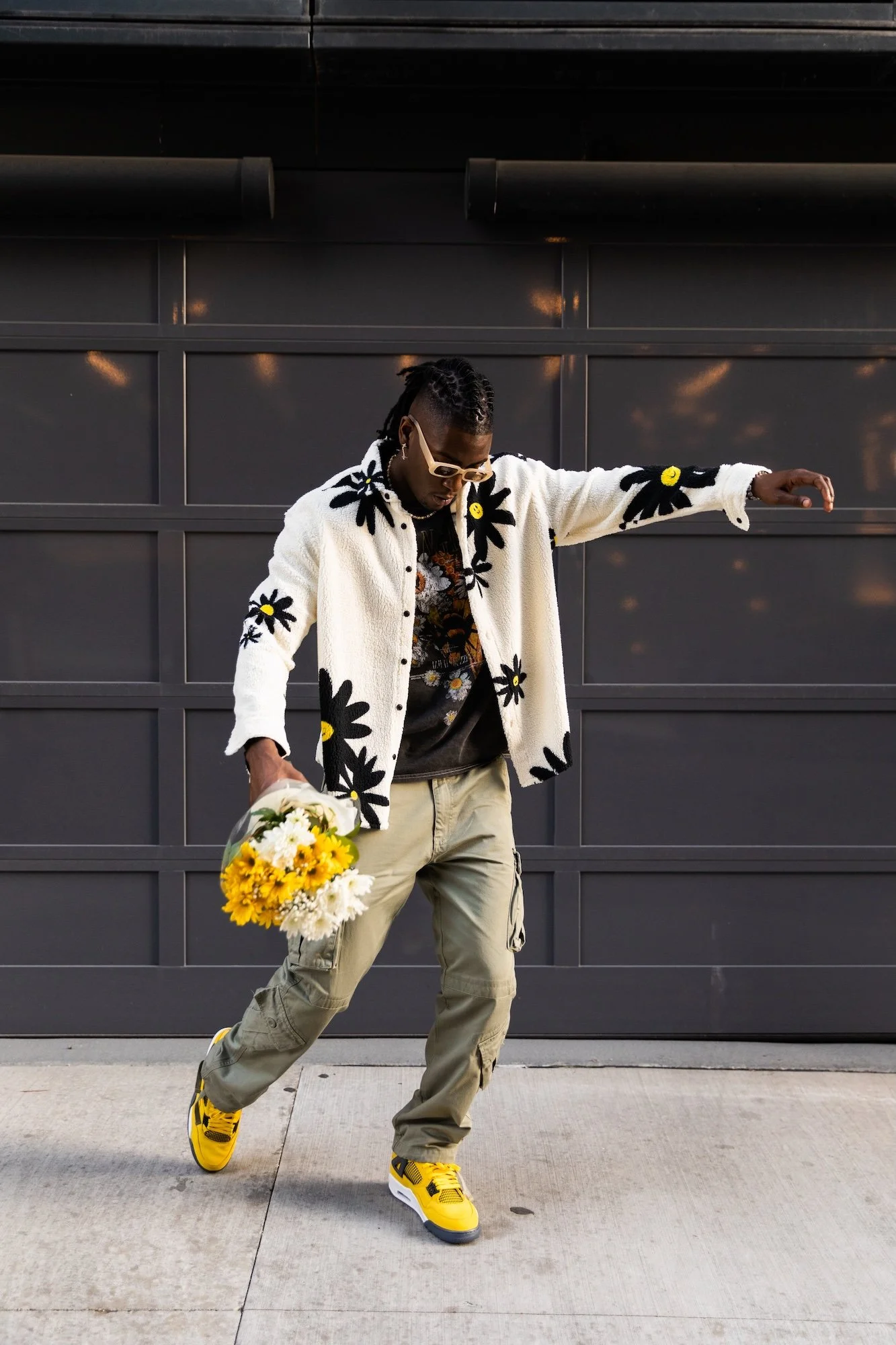 Person carrying a bouquet of sunflowers and wearing a floral jacket, beige cargo pants, and yellow sneakers, standing in front of a dark garage door.
