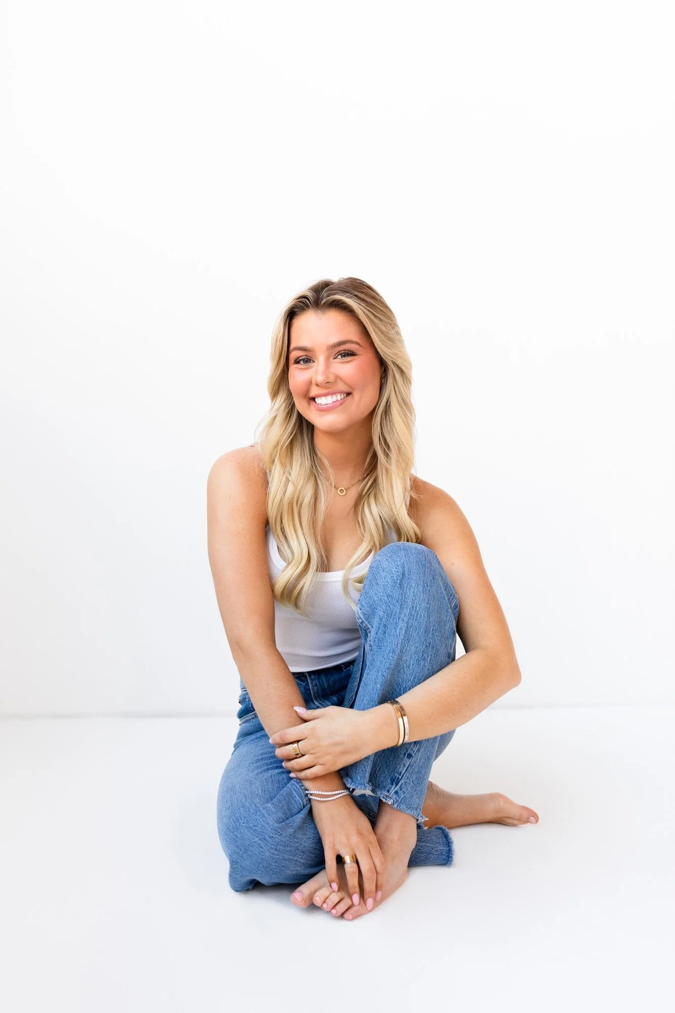 A smiling young woman with long blonde hair, sitting cross-legged on a white floor against a white background, wearing a white tank top and blue jeans.