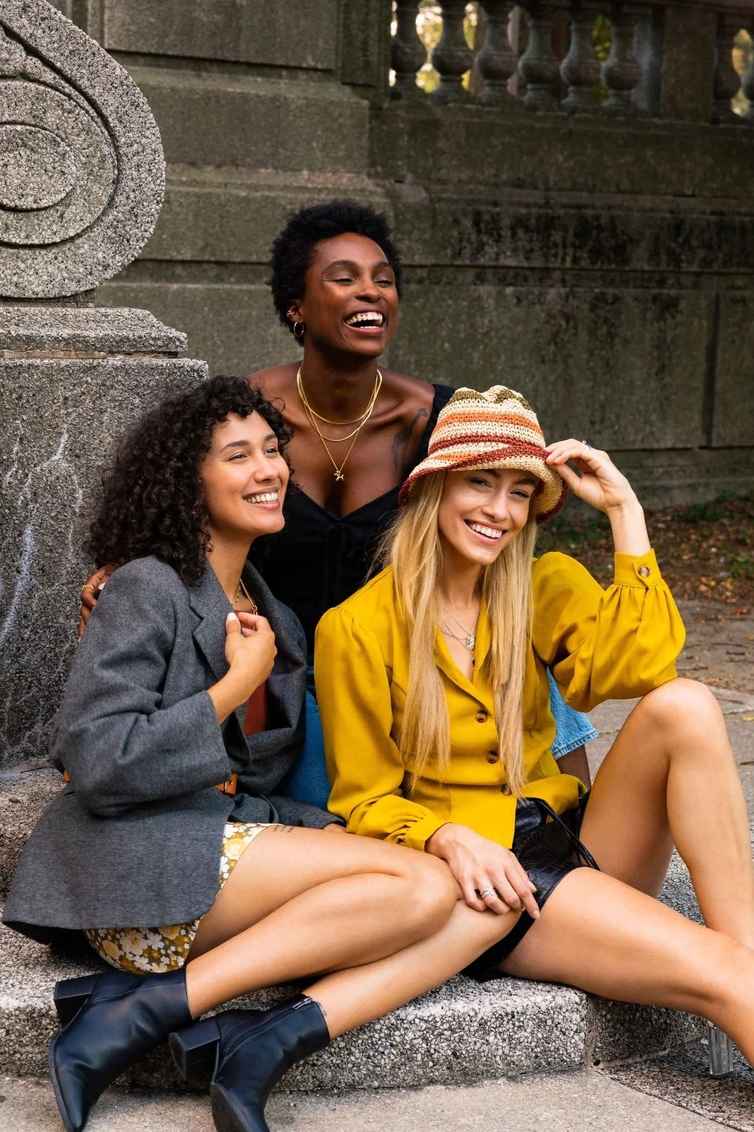 Three women sitting on a stone ledge outdoors, smiling and laughing together. One woman is standing behind them, also smiling. One woman wears a yellow shirt and a hat, another wears a grey blazer and floral shorts, and the third wears a black top and necklace. They appear happy and relaxed.