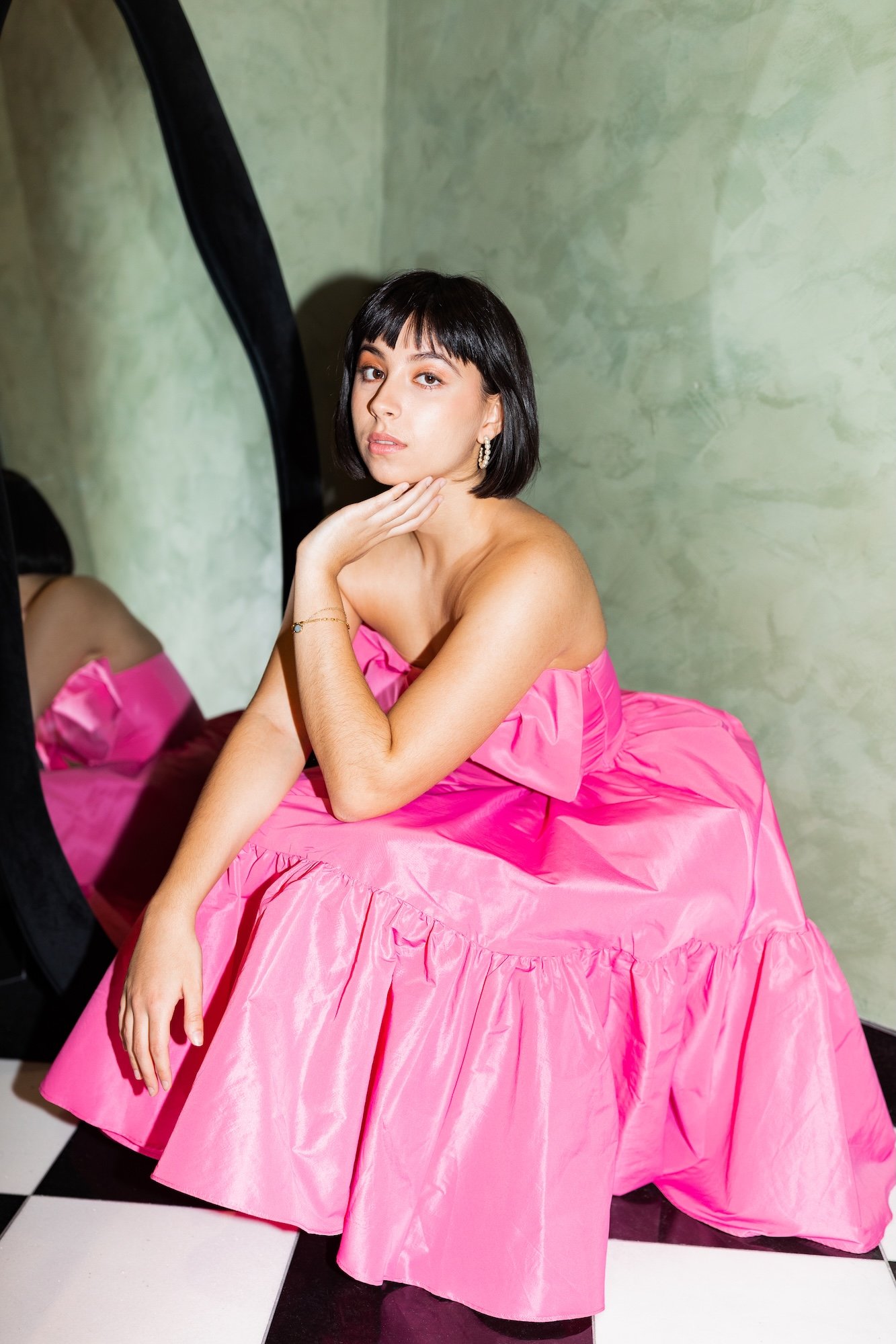 A woman with short black hair and earrings poses in front of a mirror, wearing a pink strapless dress with a ruffled skirt, sitting on a black and white checkered floor.
