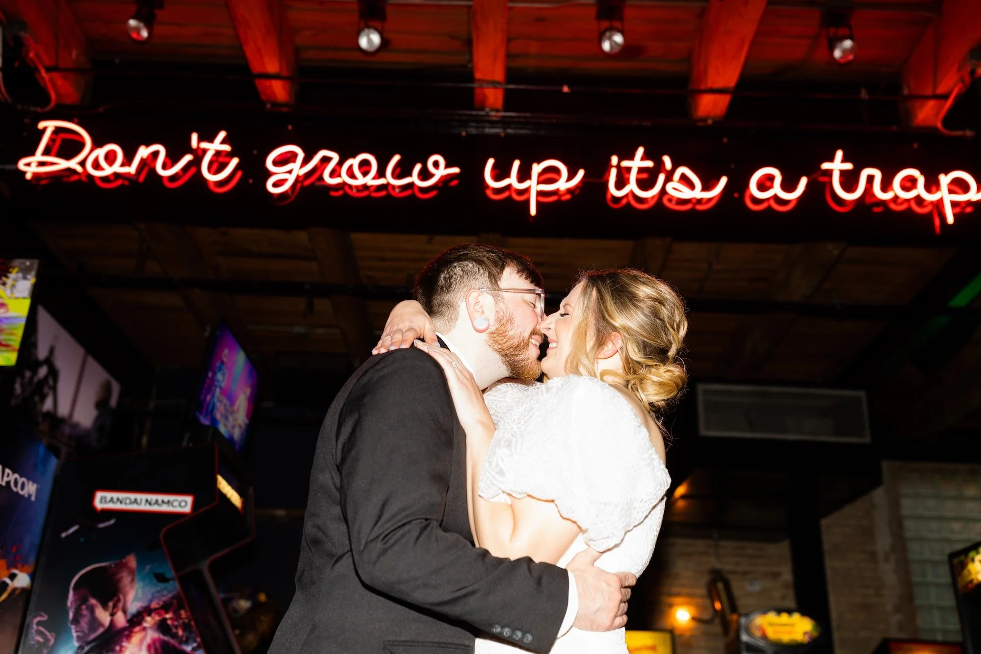 A couple dressed in wedding attire, embracing and touching noses, in a dimly lit venue with a neon sign that reads 'Don't grow up it's a trap' in the background.
