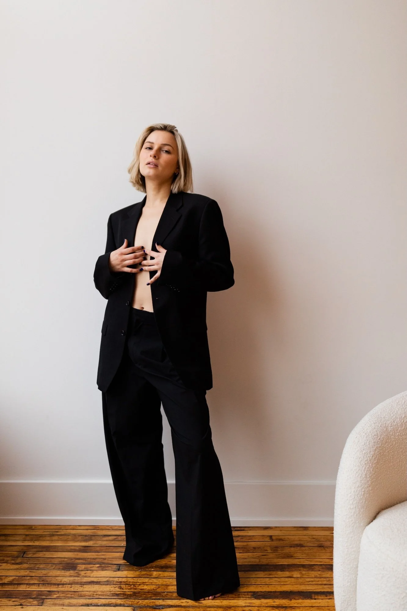 A woman in a black suit with no shirt underneath, standing against a plain white wall. She has shoulder-length blonde hair and is holding her chest with both hands.