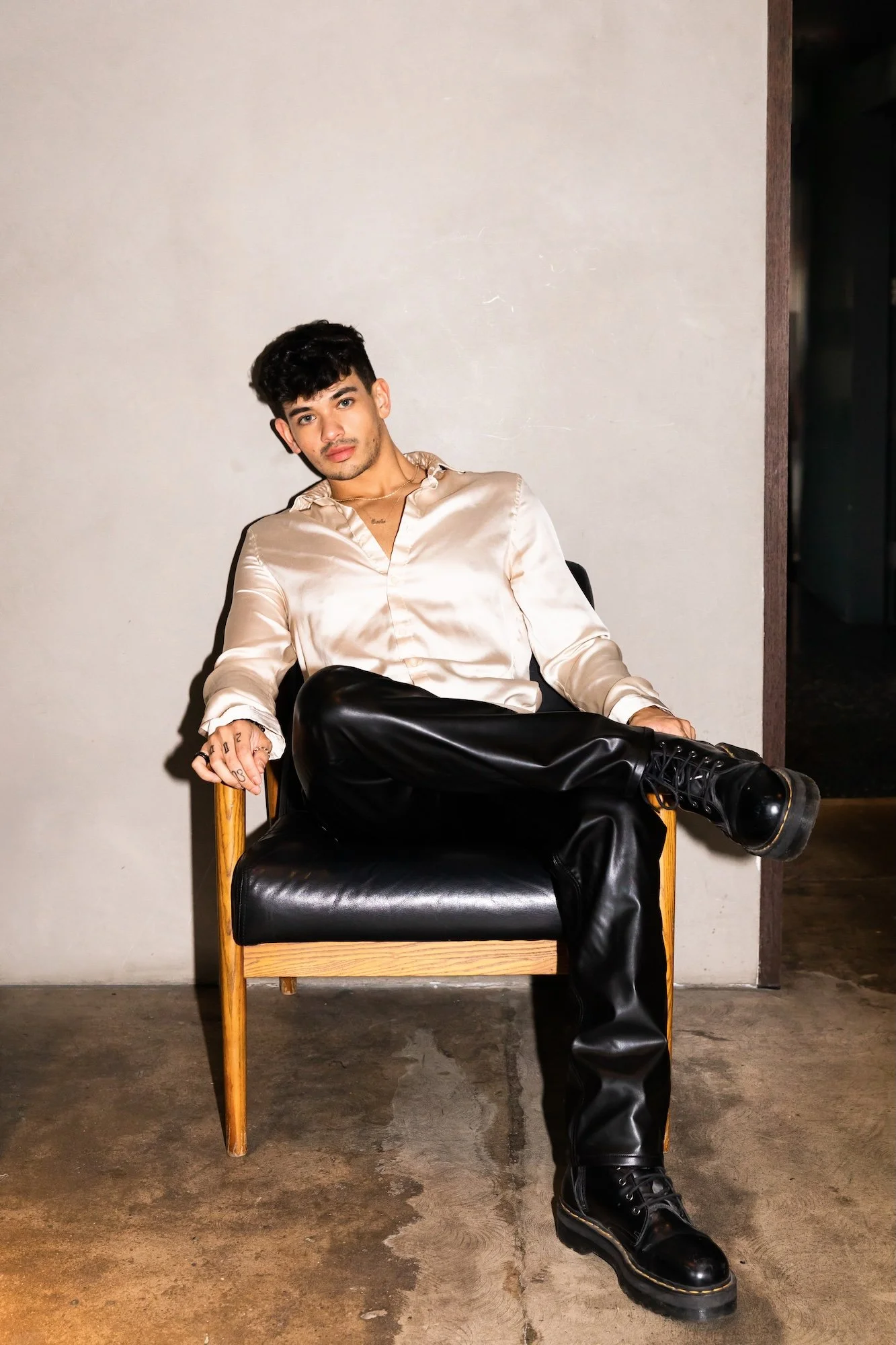 A young man with dark hair and a mustache, wearing a shiny beige satin shirt and black leather pants, sitting cross-legged in a wooden armchair with black leather cushion, against a plain light-colored wall.