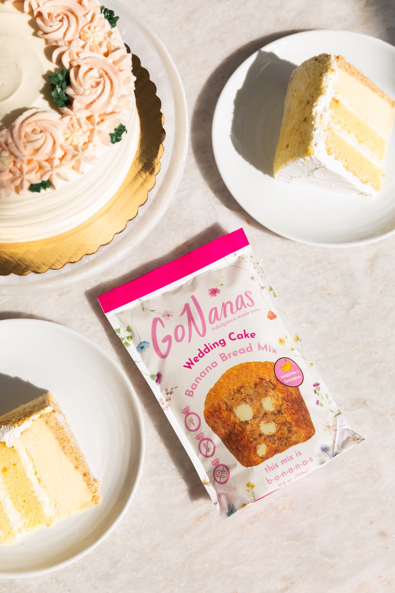 A wedding cake with pink roses and green leaves on top, two slices of white cake on white plates, and a banana cake mix packet labeled 'GoManas Wedding Cake Banana Bread Mix' on a light-colored surface.
