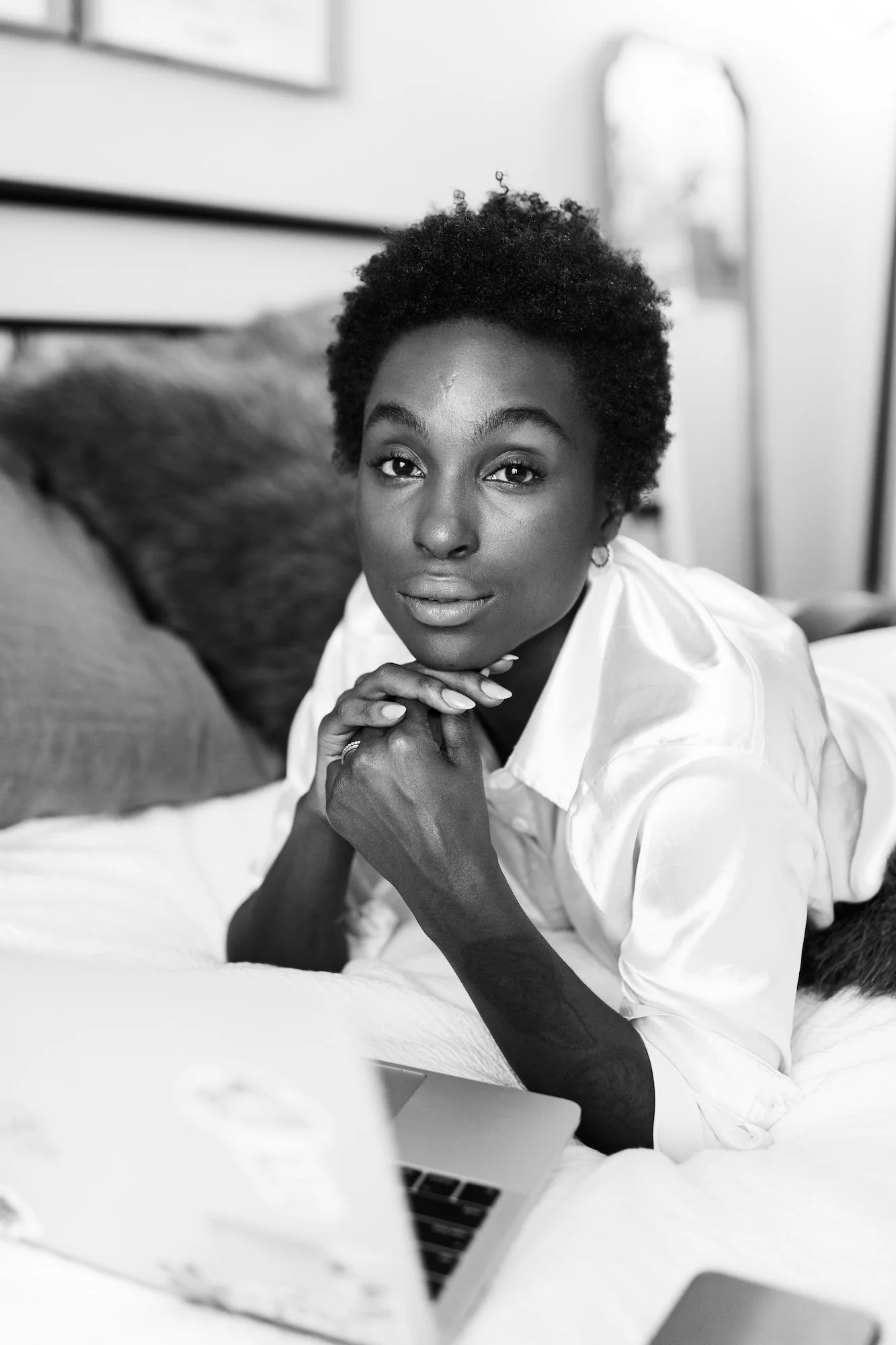 A woman with short curly hair lying on a bed, resting her chin on her hands, and looking at the camera. There is a laptop in front of her.