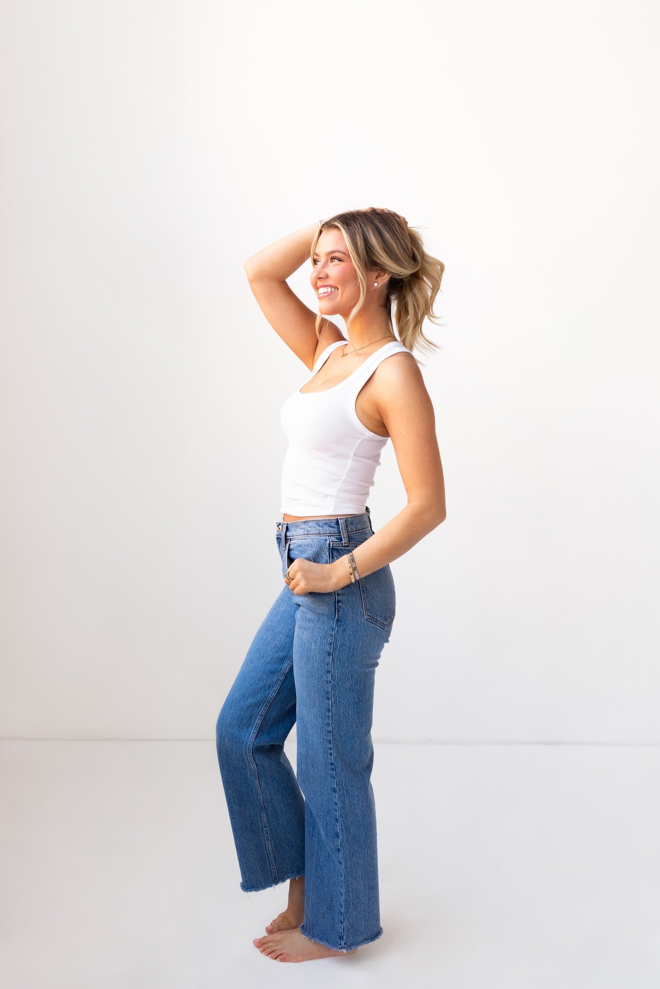 A young woman with blonde hair wearing a white tank top and blue jeans, smiling and standing barefoot against a plain white background.