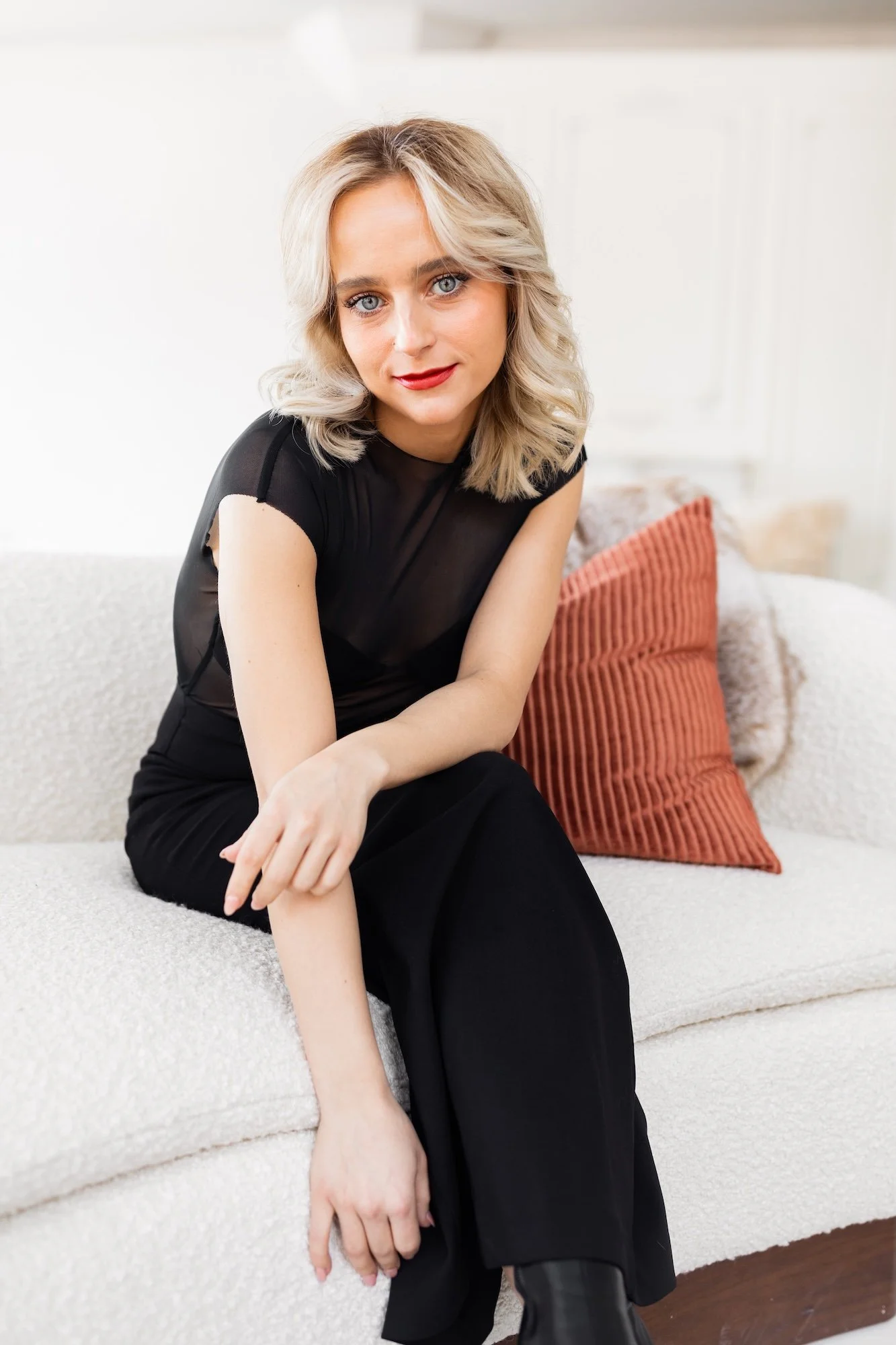 A woman with blonde, shoulder-length, wavy hair and blue eyes sitting on a cream-colored sofa with a rust-colored pillow behind her, wearing a sheer black top and black pants.