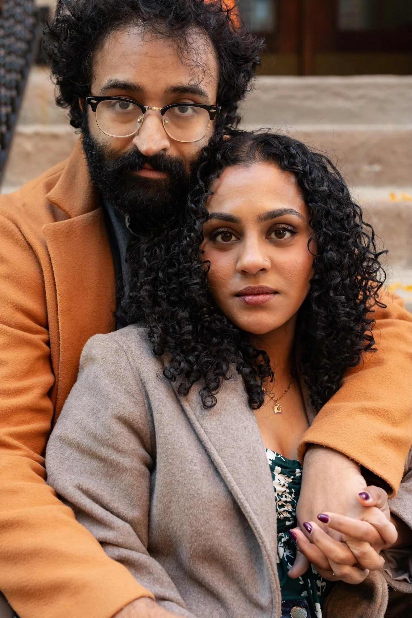 A man and woman sit closely together on outdoor steps, with the man's arms around the woman. The man has curly hair, a beard, and glasses, and wears a brown coat. The woman has long curly hair, brown eyes, and wears a gray blazer with a patterned top and a necklace. The background shows stone steps and a building.