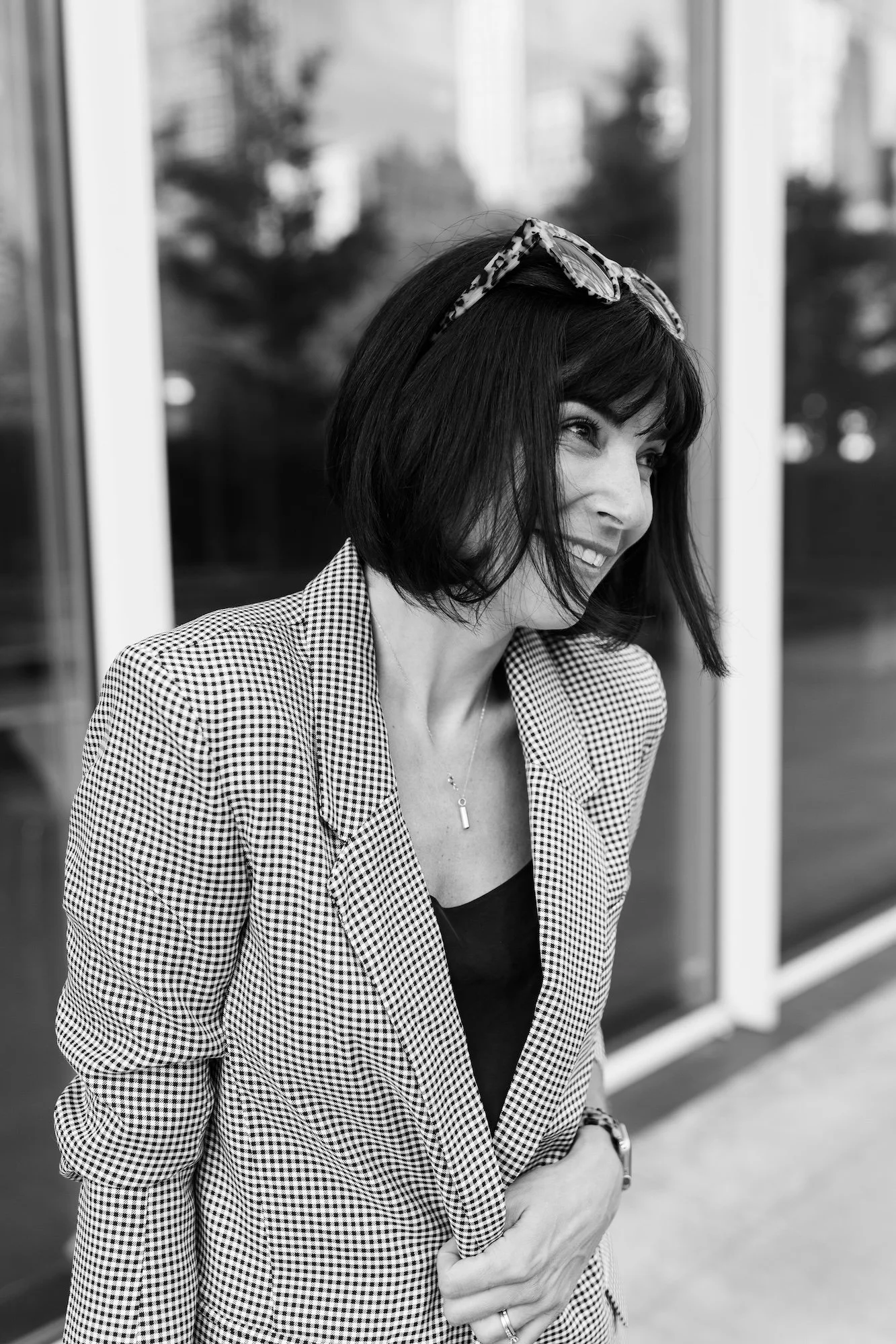 A woman with short dark hair wearing sunglasses on her head, a checkered blazer, a black top, a necklace, a ring, and a watch, smiling while standing outside near a glass building.