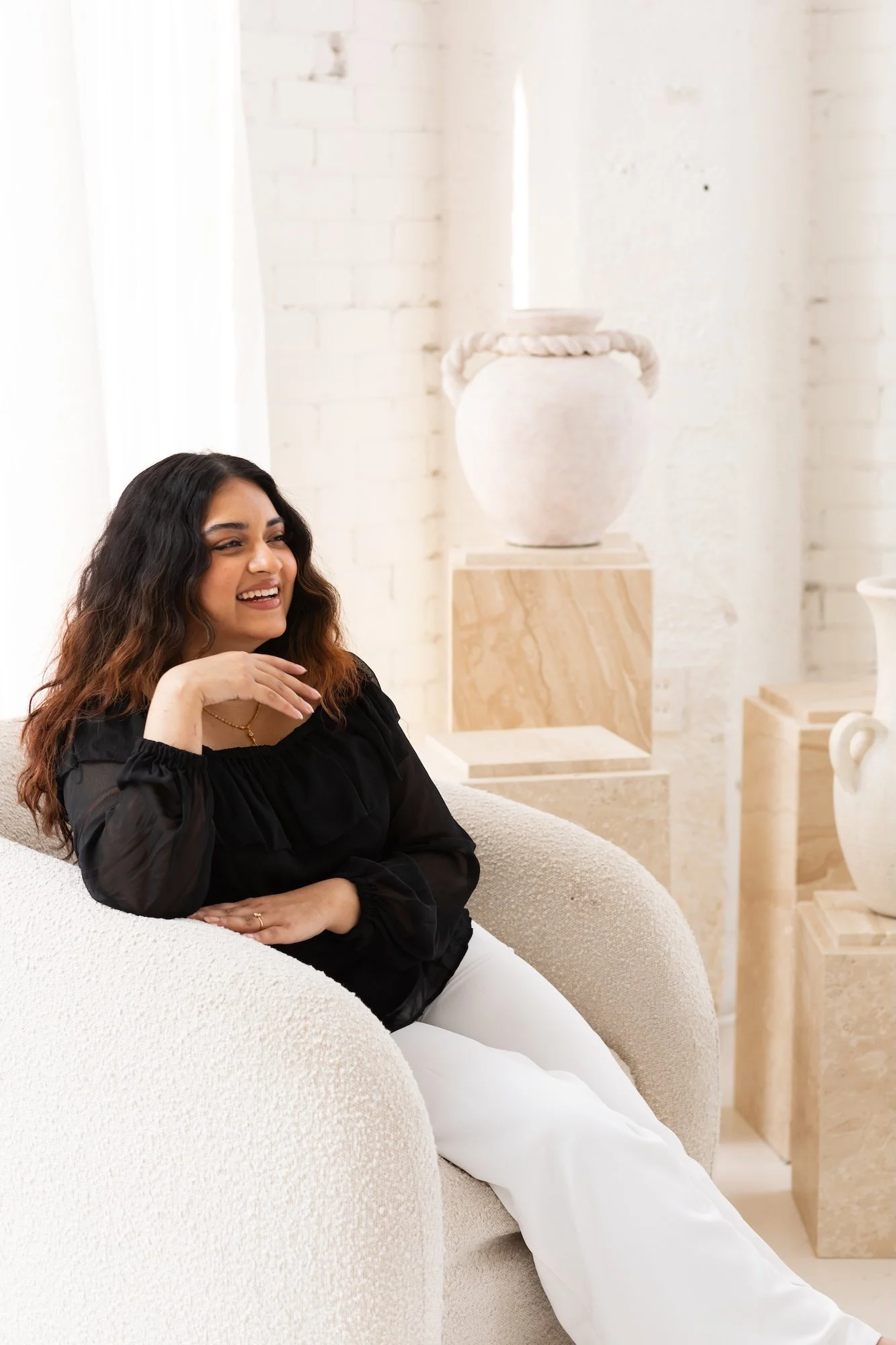 A woman with long dark wavy hair, smiling, sitting on a light-colored textured armchair in a bright room with white brick walls and decorative vases in the background.