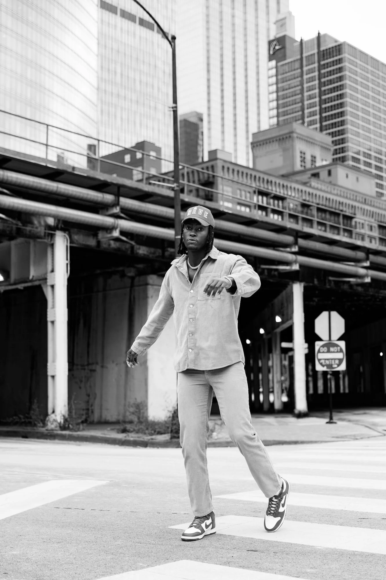 A man crossing the street in an urban area with tall buildings and an overpass above, black and white photo.