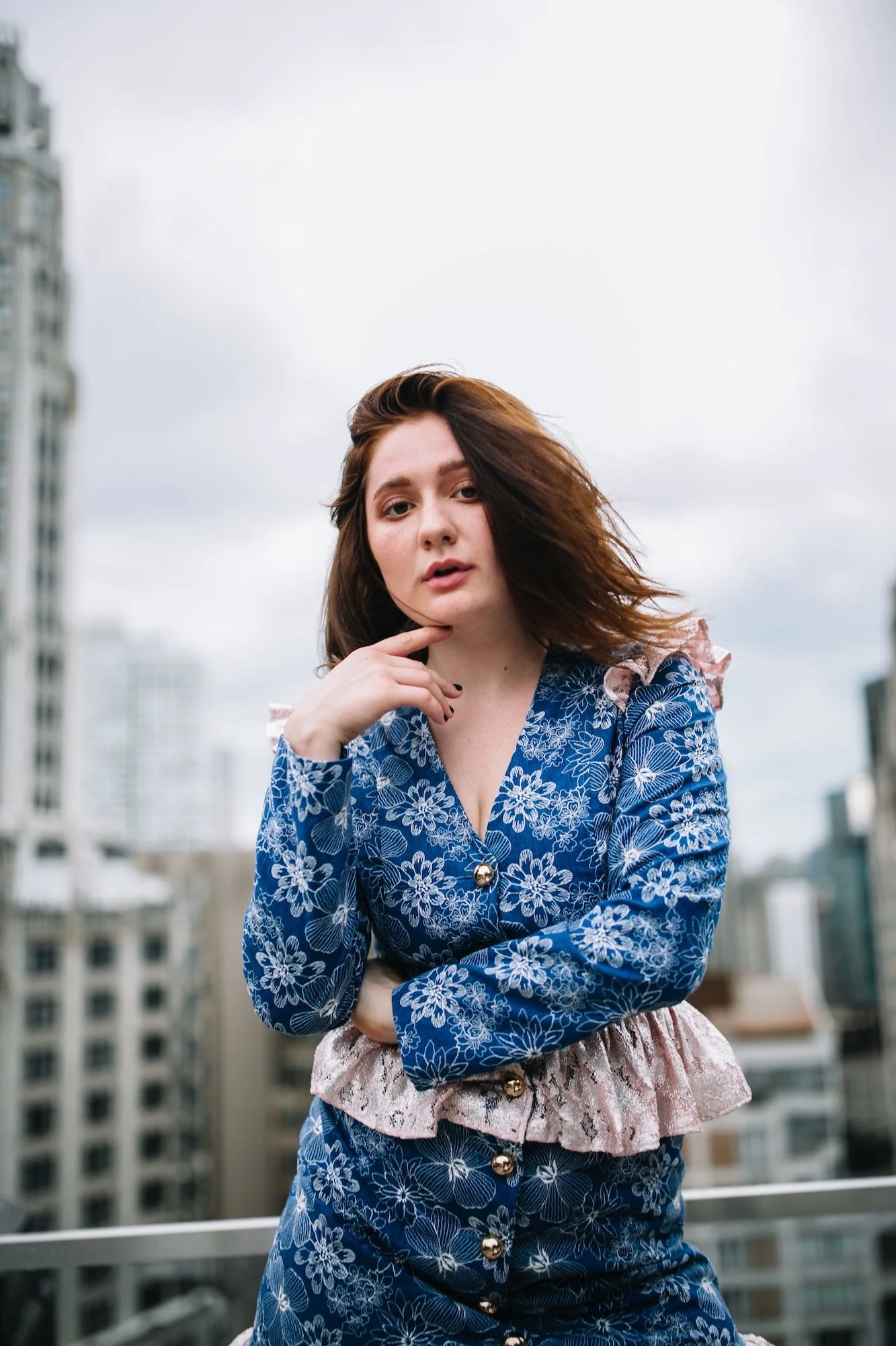 A young woman with shoulder-length brown hair, wearing a blue floral patterned dress with ruffled pink accents on the sleeves and waist, standing on a city rooftop with tall buildings in the background under a cloudy sky.