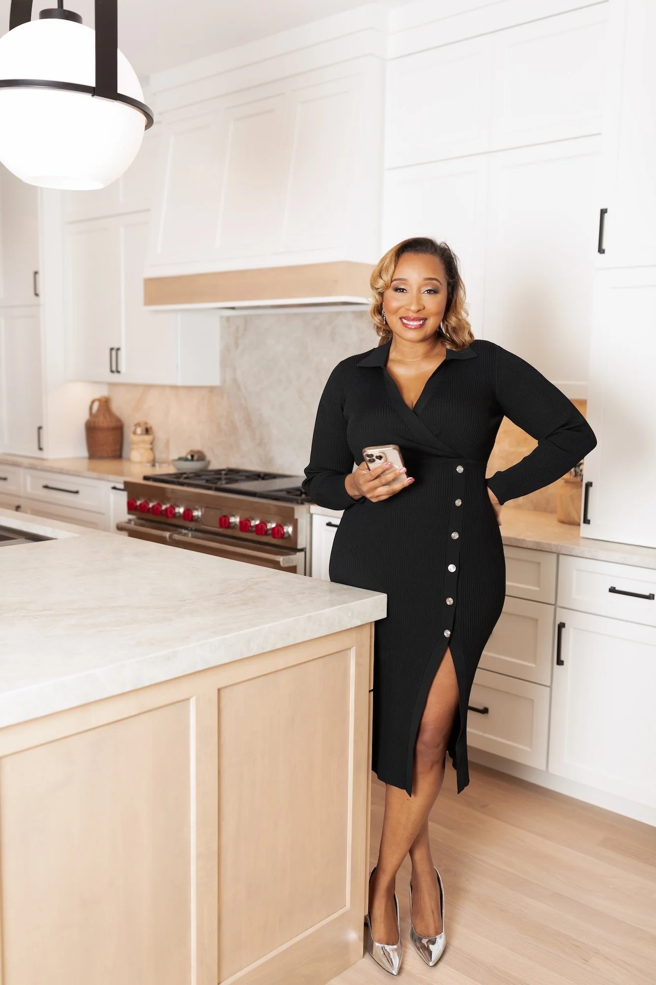 A woman in a black dress with buttons and a slit, standing in a modern kitchen, smiling and holding a smartphone.