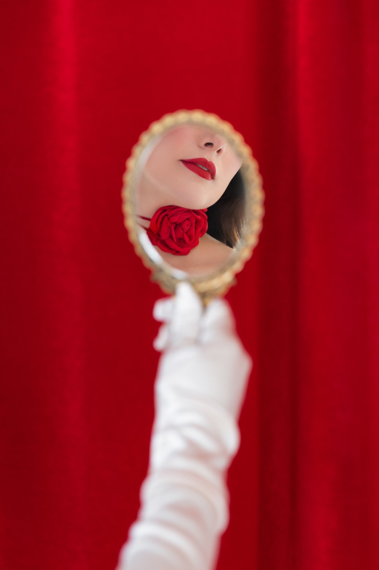 A woman holding a small ornate mirror reflecting her face, with red lipstick and a red rose necklace, against a red background.