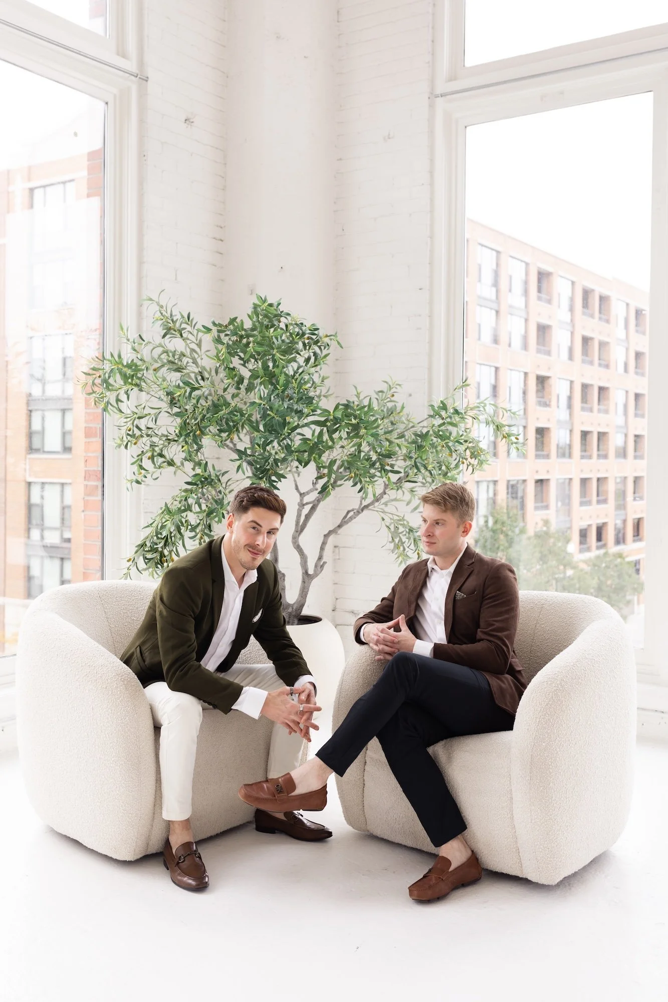 Two men sitting in modern beige armchairs with a large potted plant behind them, in front of tall windows with city buildings outside.
