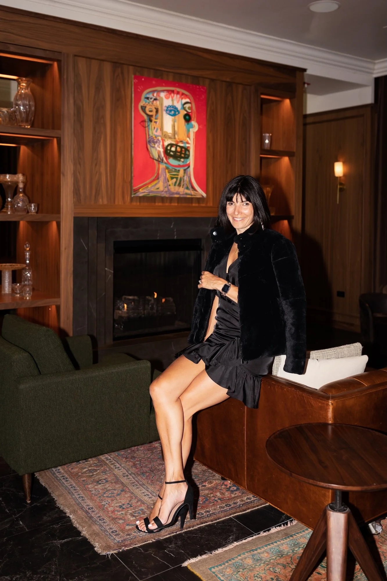 A woman with black hair wearing a black dress, black heels, and a black jacket, sitting on the back of a brown leather sofa in a living room with a fireplace, wooden walls, and colorful abstract artwork on the wall.