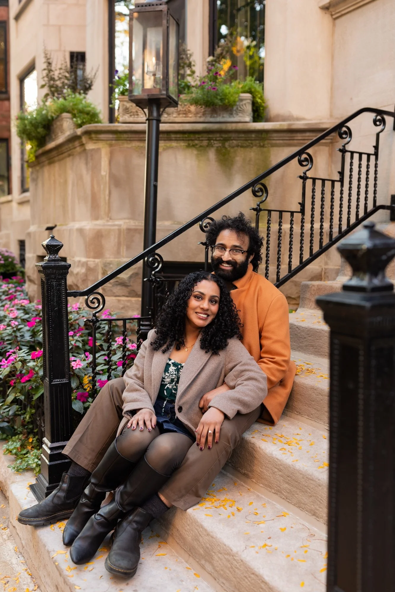 A smiling couple sitting on front steps of a house, with one person sitting on the other's lap. The woman has curly black hair, is wearing a beige coat and black boots, and the man has curly hair and glasses, is wearing an orange jacket. The steps are surrounded by pink flowers and a black wrought iron railing, with a decorative lantern above.