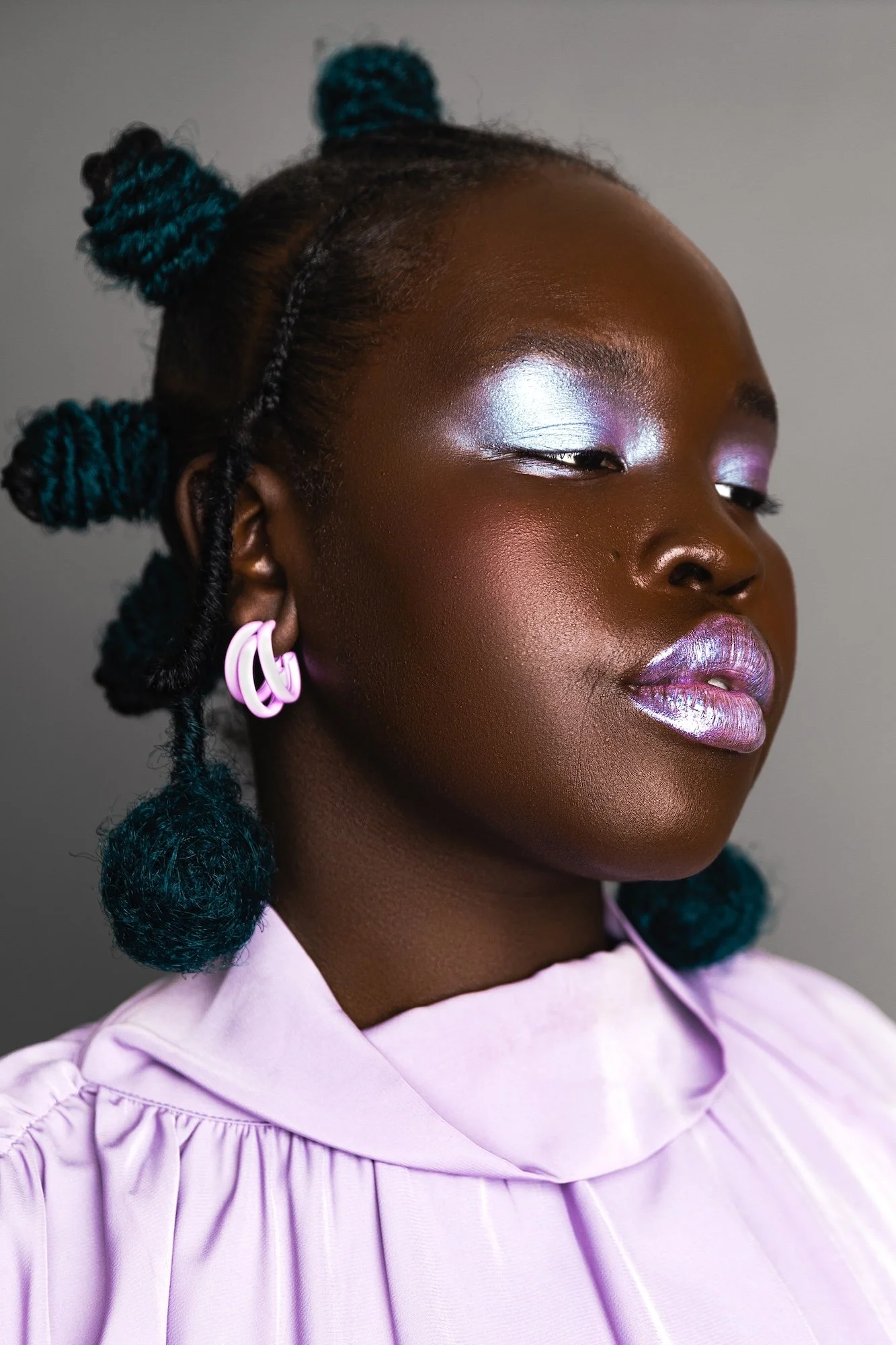 Close-up of a woman with dark skin, styled in bantu knots with teal accents, wearing pastel pink earrings and shimmery pastel makeup, dressed in a light pink ruffled top.