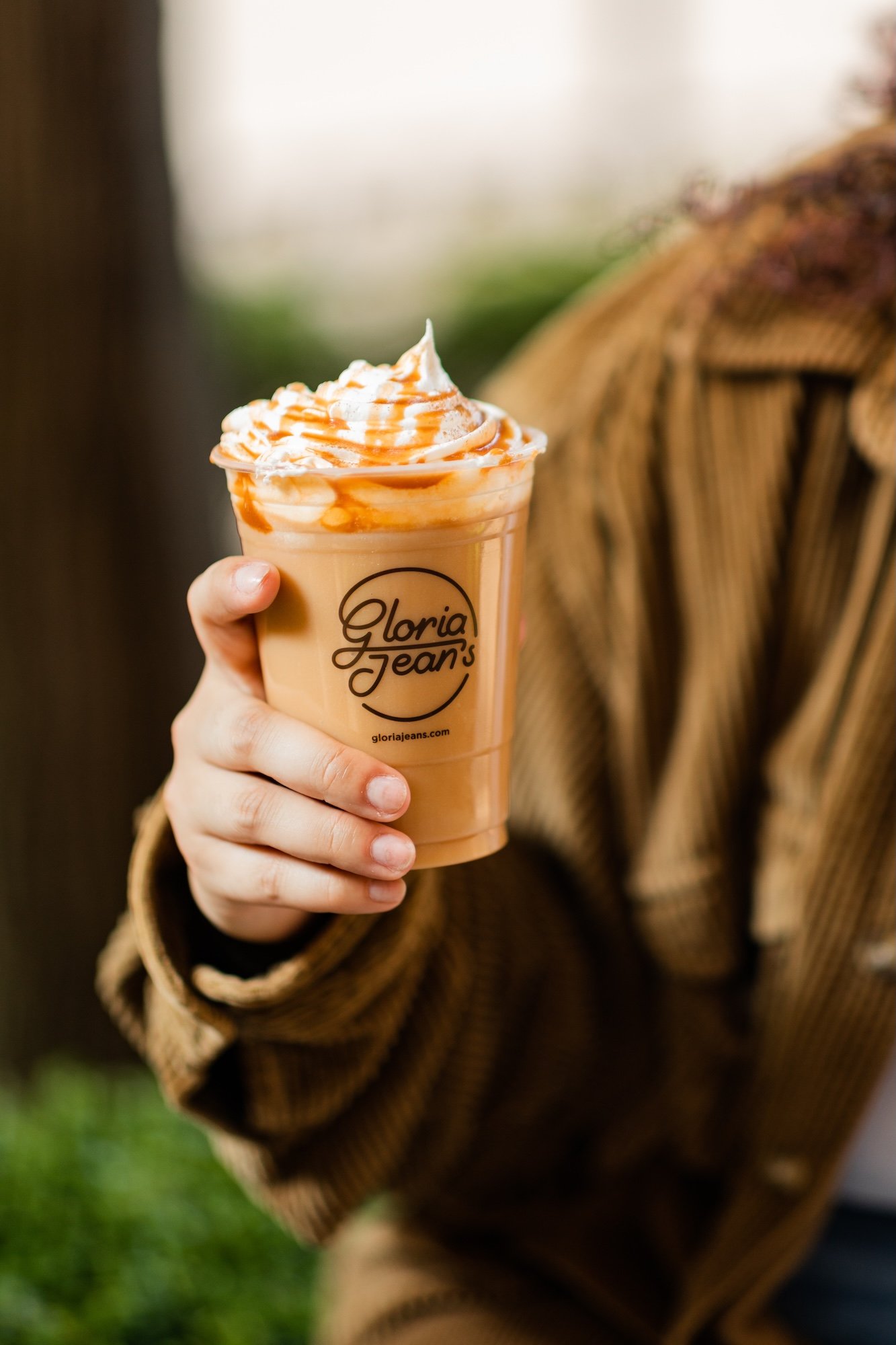 A person holding a caramel-flavored iced coffee with whipped cream and caramel drizzle, in a clear plastic cup with a Gloria Jean's logo.
