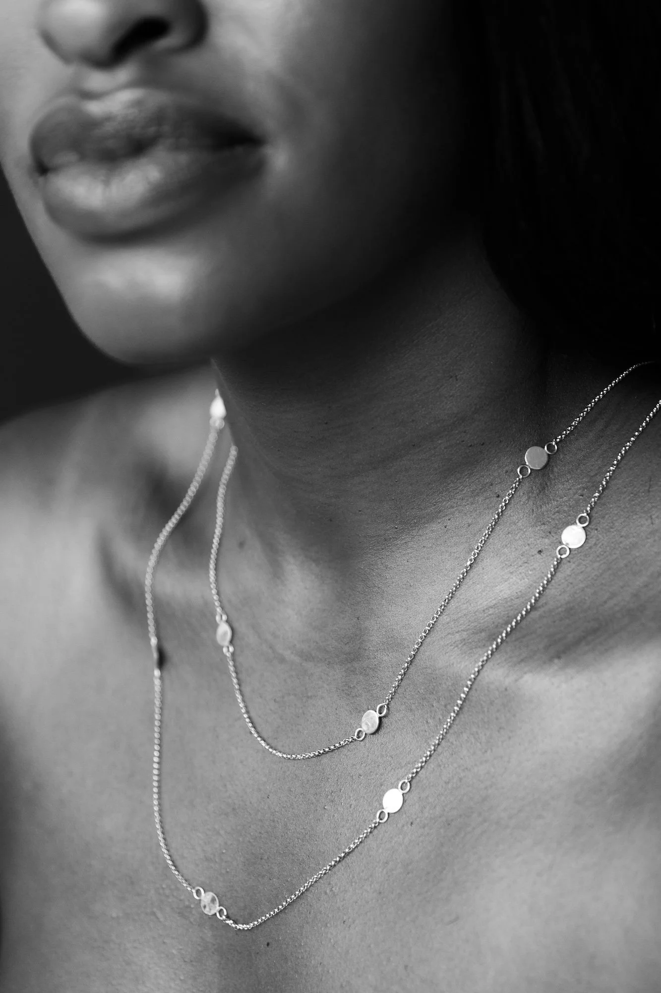 Close-up black and white photo of a woman's neck and lips, wearing layered necklaces with circular pendants.