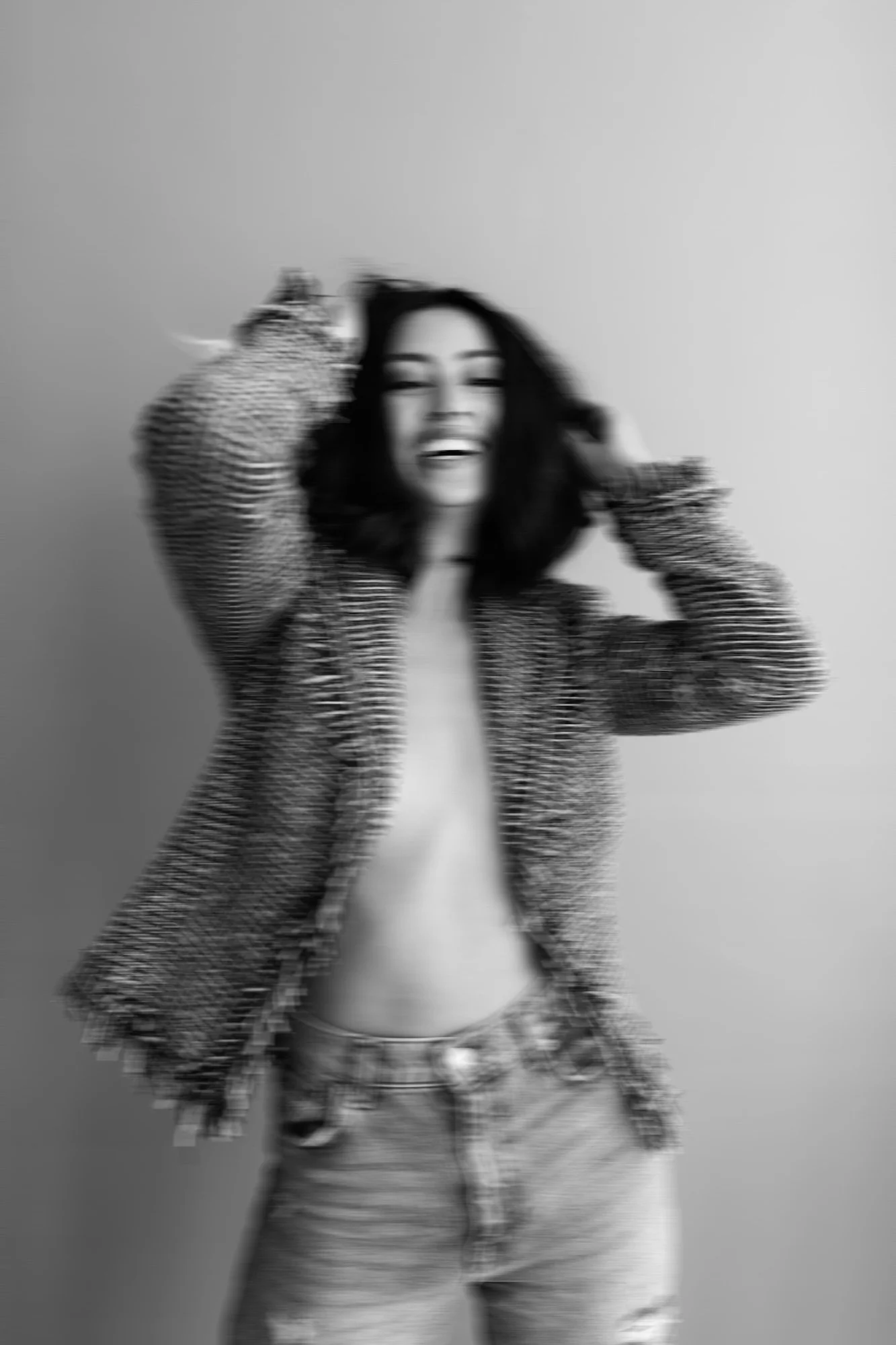 Blurry black-and-white photo of a young woman laughing, wearing a striped jacket and jeans, posing against a plain wall.